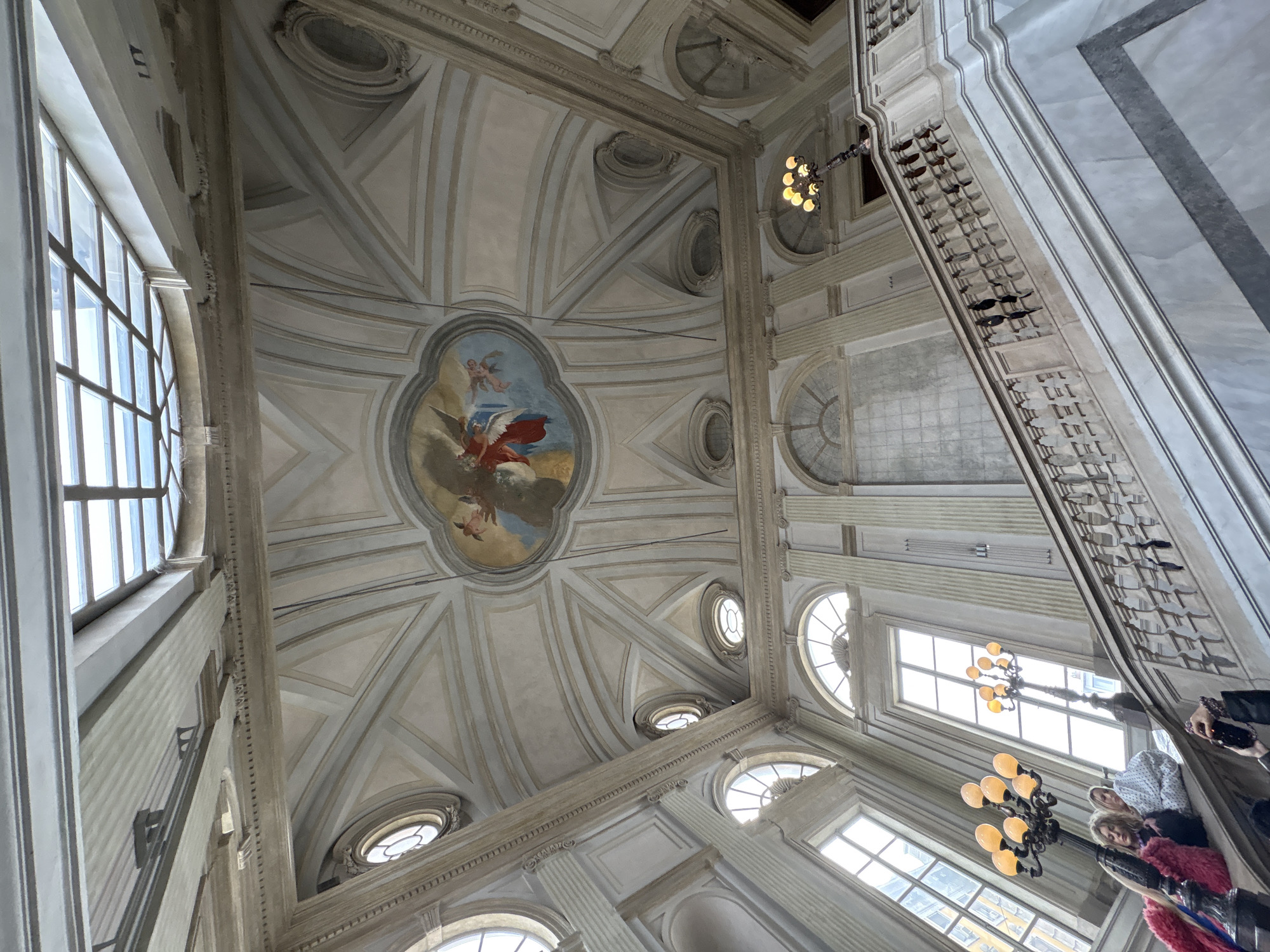 The image shows an ornate interior view looking upwards, possibly the ceiling of a building in Milan, Italy, based on the architectural style. The ceiling is primarily off-white with intricate geometric patterns and a central painting. The painting features figures in what seems to be a classical or religious scene. There are several windows providing natural light. Two chandeliers with multiple bulb arms are visible. In the lower right corner, people are partially visible, likely looking at the architecture, and taking photographs. A staircase is seen with ornate railings.