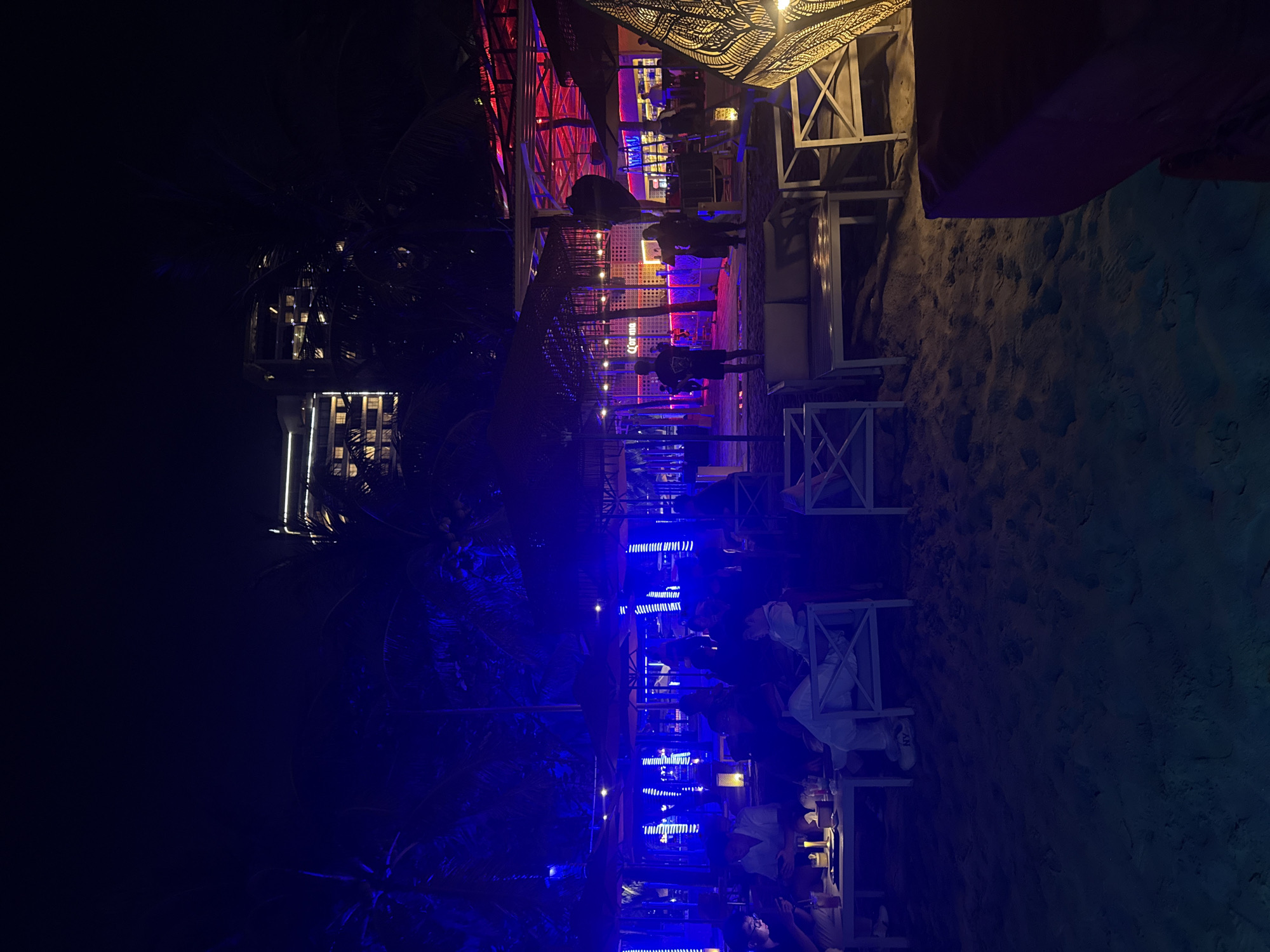 The image shows an outdoor nighttime scene at what appears to be a beach bar or restaurant in an unknown location. The foreground features tables with seated patrons, illuminated with blue lighting. Beyond this area, the image suggests a structure with red and purple lighting, likely part of the bar or restaurant. Additional details include palm trees on the left side of the frame and sandy ground in the foreground and to the right. Some individuals can be seen walking or standing near the brightly lit structure. Overall, the image depicts an active nightlife setting.