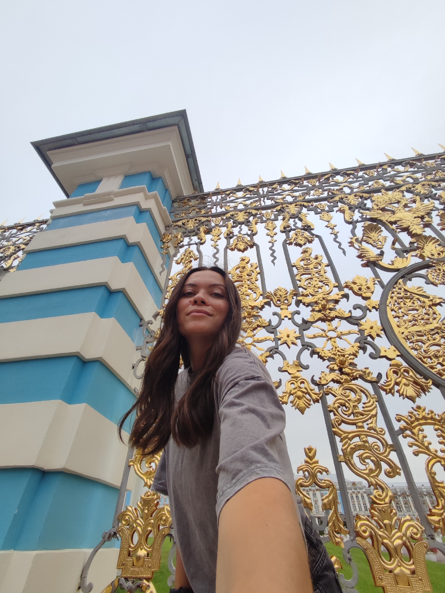 A low-angle, wide shot captures a young woman with long, dark hair taking a selfie in front of a large, ornate golden gate and a blue and white striped pillar. The woman is wearing a grey t-shirt and is positioned in the lower right quadrant of the frame, her face angled slightly upwards towards the camera. The golden gate is intricately detailed with swirling patterns and what appear to be animal motifs, set against a pale sky. To the left of the gate is a monumental pillar with alternating horizontal bands of bright blue and white. In the background, beyond the gate, a distant view of a grand white building with a neoclassical facade is visible. The scene is identifiable as the Catherine Palace in Pushkin, Russia, given the distinctive architecture of the palace and the elaborate golden gate. The lighting is soft and diffused, suggesting an overcast day.