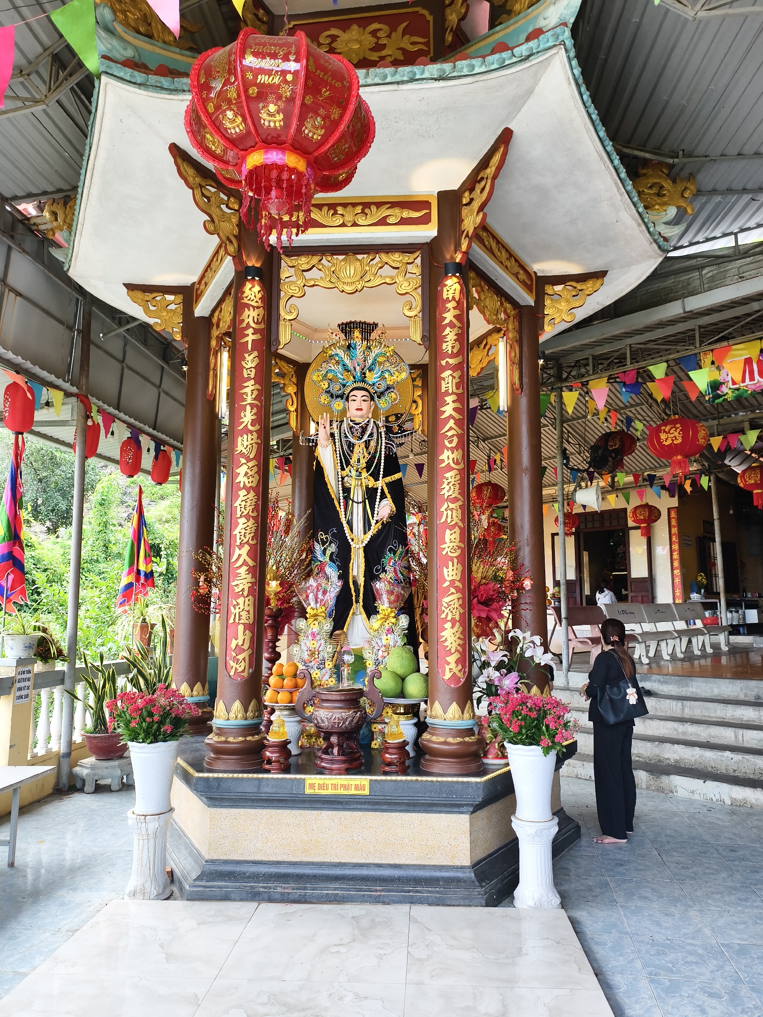 Captured in Tay Nha Trang Ward, Vietnam, a religious shrine dominates the scene, housing a statue adorned in black and gold attire. A red lantern hangs above the shrine, adding to the solemn atmosphere. Columns are embellished with inscriptions in a gold color, and offerings of fruits and flowers are placed on the altar in front of the statue. A woman in black pants and a top, facing the shrine, is present in the background, apparently in prayer. Colorful flags are seen hanging nearby, and potted plants stand on either side of the shrine's base, enhancing the spiritual ambiance of the location.