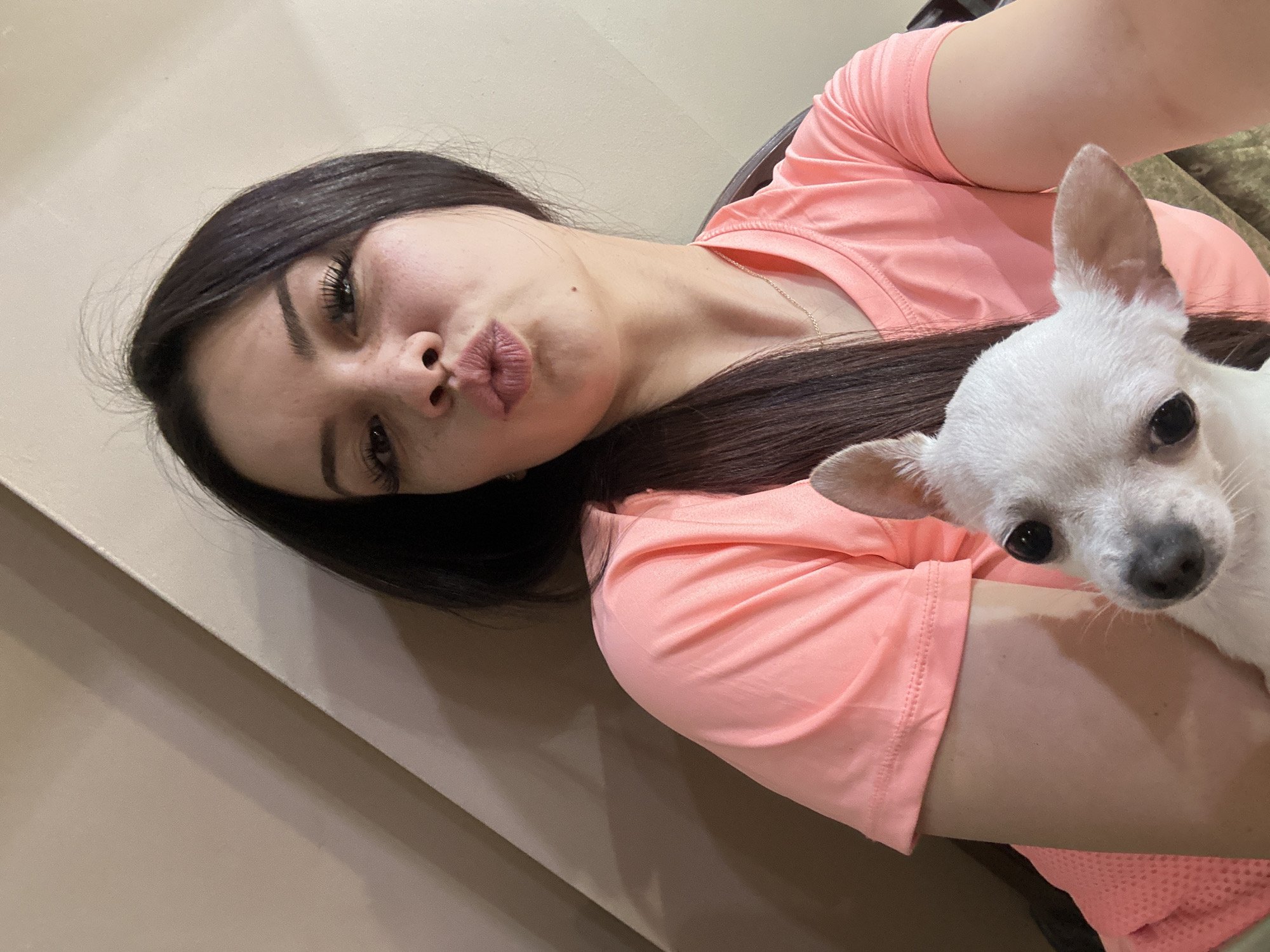 A young woman with dark hair and freckles is taking a selfie with a white chihuahua. She is wearing a coral-colored t-shirt and is making a kissing face. The dog is held in her arms, and its head is partially visible in the lower left corner of the image. The background is a neutral-colored wall. The image appears to have been taken indoors. The location is not specified.