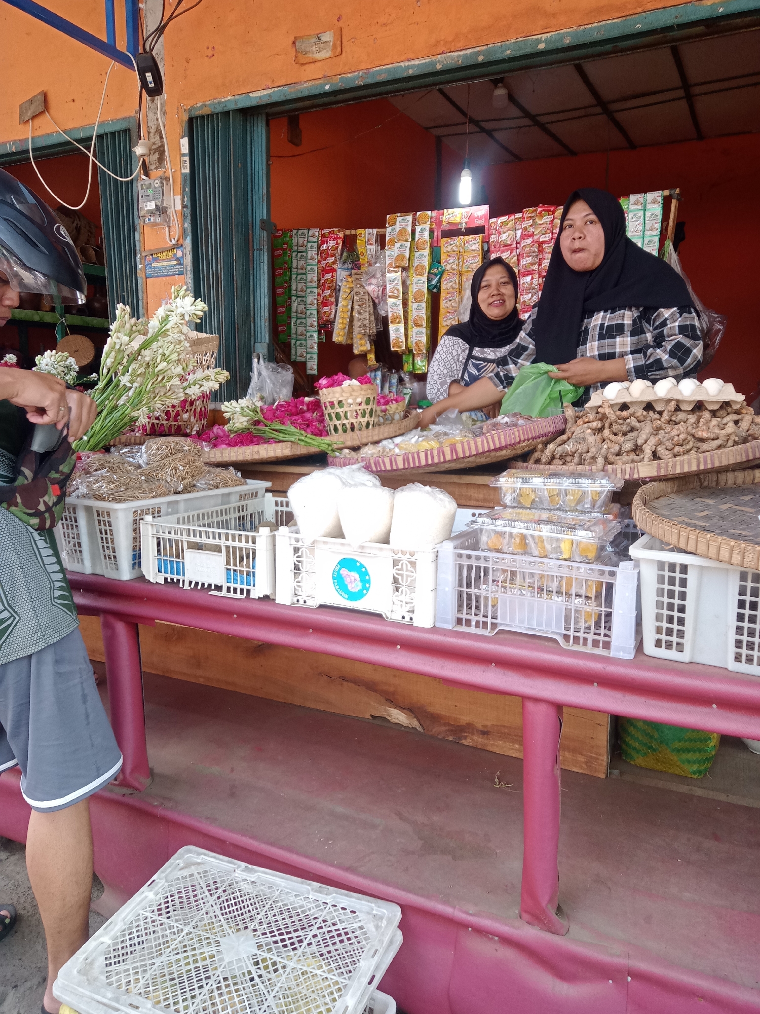 In Borobudur, Indonesia, two women stand behind a market stall displaying various goods. The stall is open-fronted with an orange-colored wall and a blue metal frame above. In the foreground, a person wearing a grey t-shirt and grey shorts is partially visible, holding a bunch of white flowers.
The stall's counter is a wooden surface covered with a red fabric. On the counter, there are several white plastic crates filled with items. One crate in the foreground contains what appear to be dry goods or produce. Other crates hold packaged goods, and one contains white, round objects, possibly coconuts or large bulbs.
Further back, on woven trays, are more items. One tray holds a large pile of ginger or turmeric roots, with a carton of eggs on top. Another tray displays dried goods tied in bundles, and a third tray holds pink flowers. Behind the women, shelves are stocked with rows of packaged snacks and food items, predominantly in red, yellow, and white packaging.
The woman on the left, wearing a headscarf and a patterned top, smiles at the camera. The woman on the right, also wearing a headscarf and a black and white checkered shirt, holds a green plastic bag. Both women are engaged in tending to the stall's merchandise. The overall scene depicts a bustling local market in Indonesia.