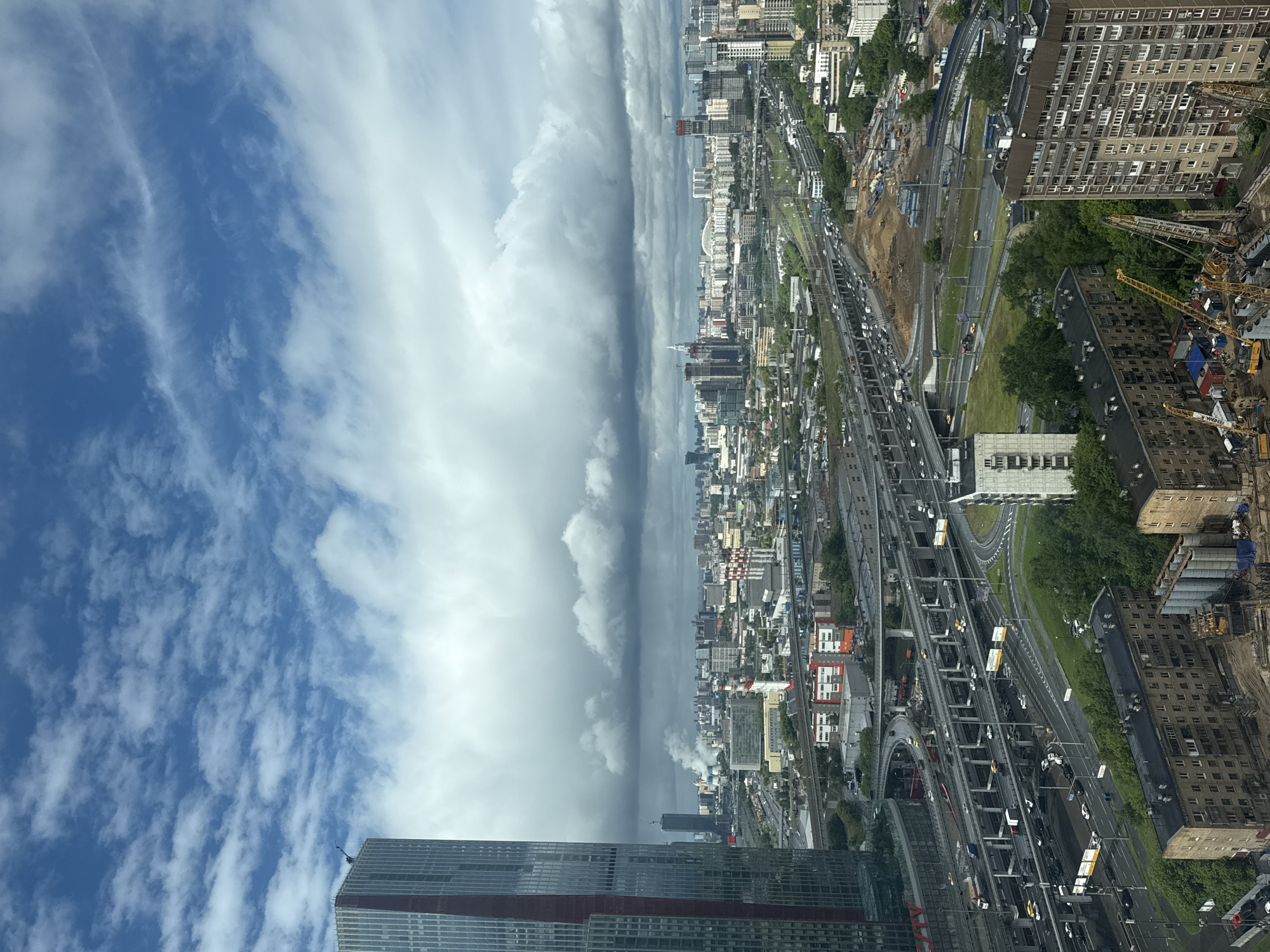 The image presents an aerial view from a high vantage point in a city, likely Khimki, Russia. A prominent skyscraper sits in the foreground. A wide band of dark, ominous storm clouds dominates the central view of the image, contrasting sharply with the brighter, blue sky and scattered cumulus clouds in the upper left. Below the clouds, a densely populated urban landscape stretches into the distance. Buildings of varying heights and architectural styles intermingle, intersected by complex road networks with heavy traffic. Construction sites with cranes are visible, indicating ongoing development. The general atmospheric conditions suggest an approaching storm.