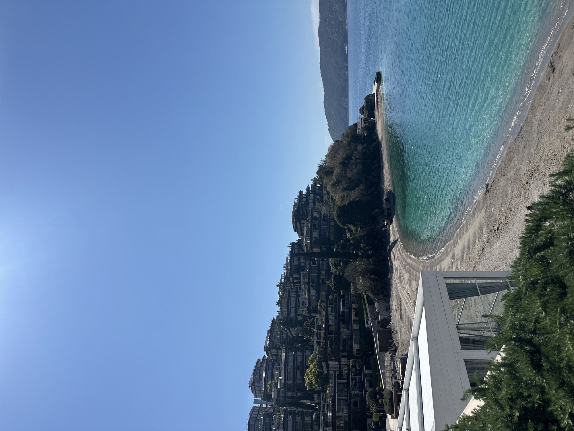 The image shows a view from the top of the Dukley Gardens complex, a luxury residential development located in Budva, Montenegro, on the Zavala peninsula. The photo captures a beach with clear turquoise waters, flanked by a mountainside covered in buildings and trees. The sky is a clear, bright blue. A white, modern architectural structure is partially visible in the foreground. There are no people visible in the image.