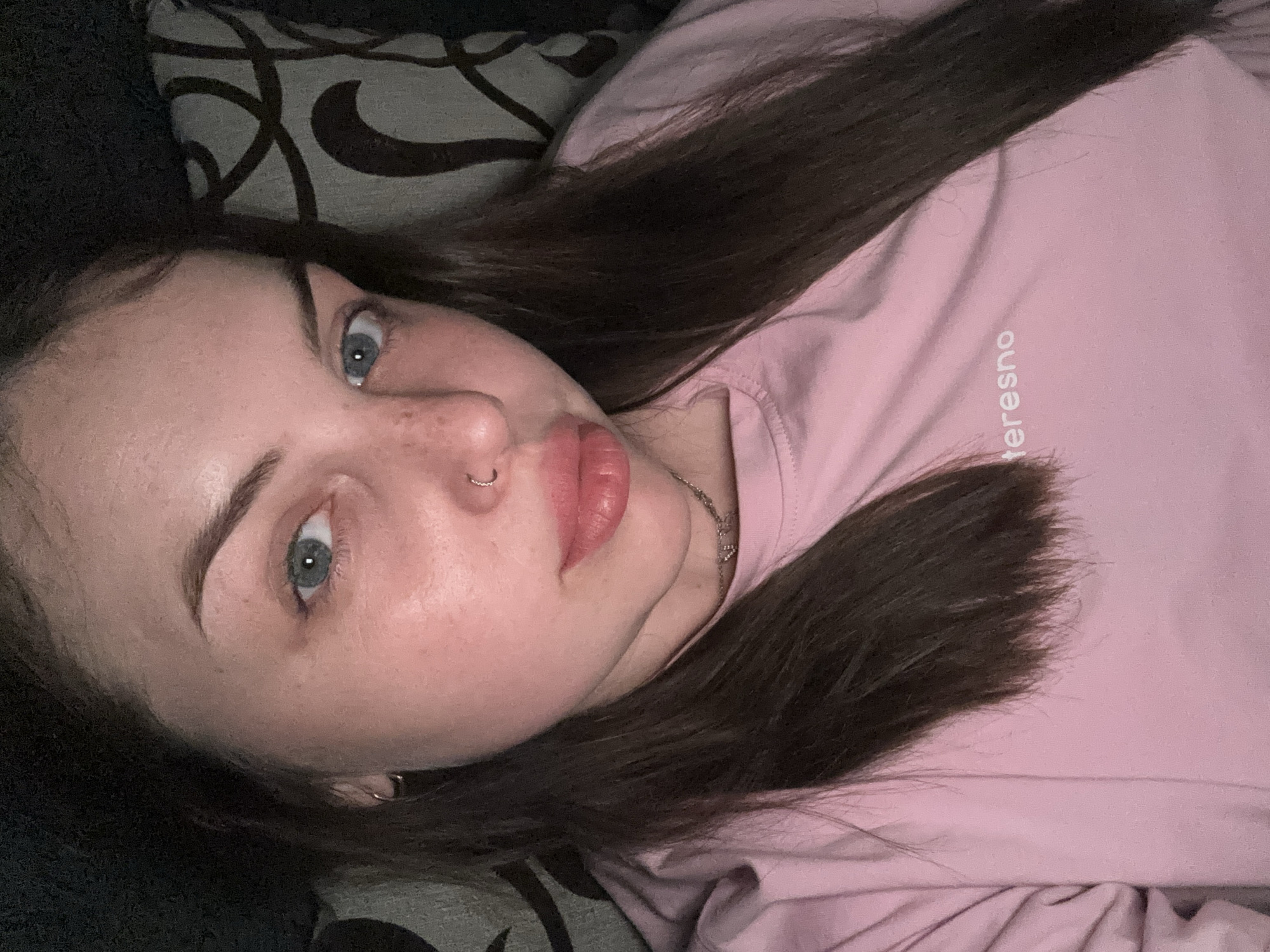 A woman is laying down, angled towards the camera. She has brown hair and blue eyes. She's wearing a light pink shirt with the word "teresno" written on it vertically. She has a nose ring and freckles on her face. She is lying on a dark-colored surface with a pillow that has a black and grey pattern. The location is not discernible from the image.