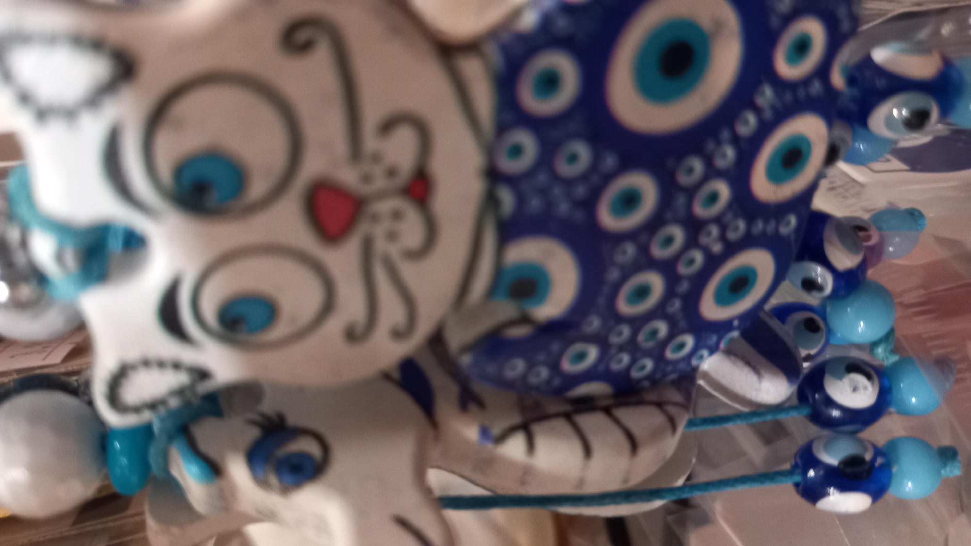 This is a close-up, blurry photograph of a decorative item featuring two cartoonish cat figures. The primary subject, in the foreground and right of center, is a white cat with large, blue eyes, black outlined features including whiskers and eyebrows, and a small red nose. This cat is wearing a blue garment adorned with a repeating pattern of concentric circles in white and blue, resembling stylized eyes. To the left and slightly behind is another, similar white figure, possibly a dragon or another cat, also with blue eyes and black facial features. Both figures are suspended by blue cords, from which hang several smaller, bead-like charms. These charms are blue and light blue, and some of them also feature the same eye-like pattern seen on the larger cat's garment. The background is out of focus, showing indistinct shelves and possibly other merchandise, suggesting the items are displayed in a shop. The location is identified as the city of Kos, Greece.