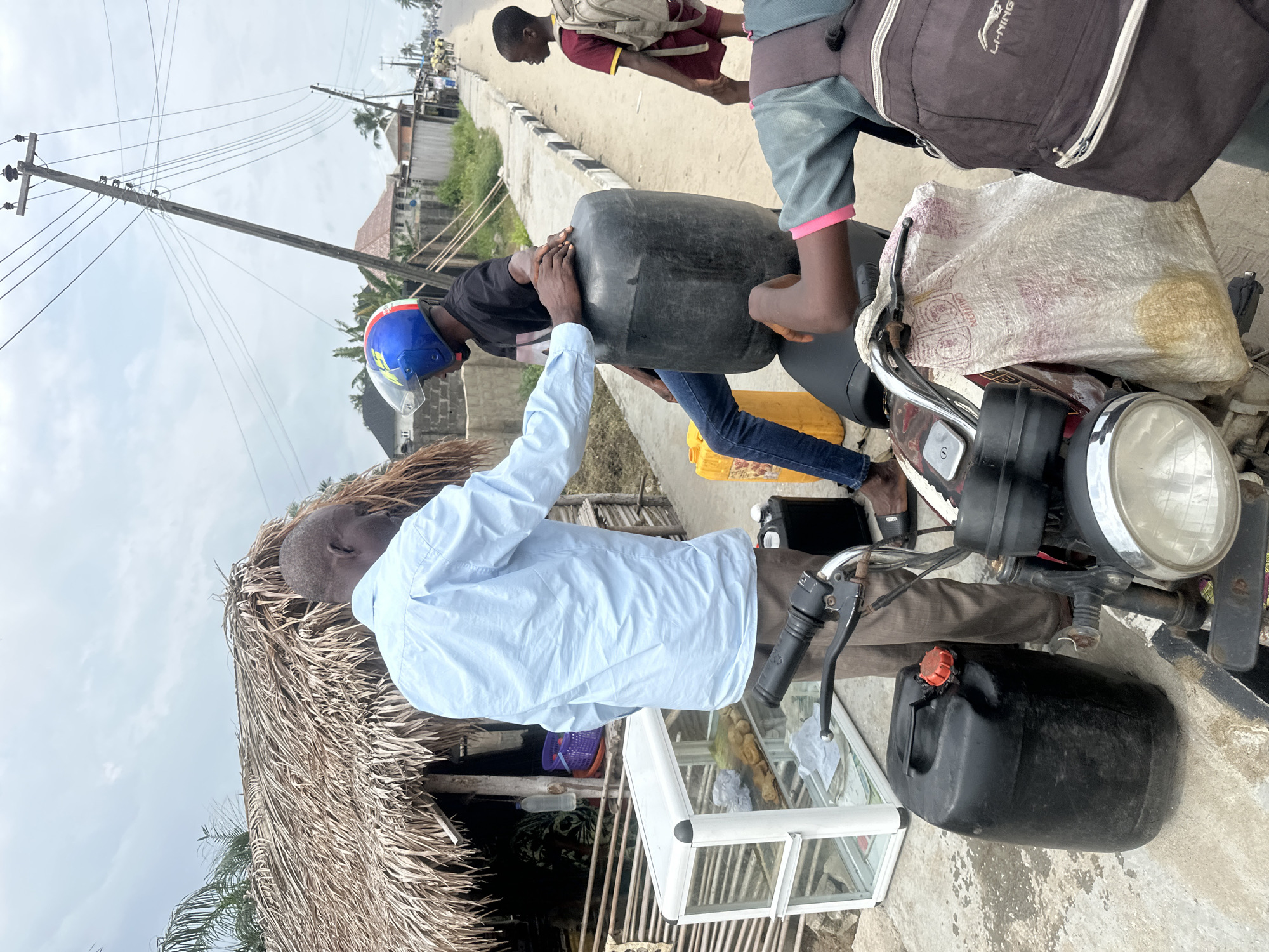This image captures a vibrant roadside scene in Badagry, Nigeria, likely during the daytime under an overcast sky. The main activity centers around a motorcycle and a transaction involving large jerrycans, suggesting a makeshift fuel or liquid vending point.
In the foreground, to the left, a man in a light blue long-sleeved shirt stands, with his back partially to the viewer. He appears to be a vendor, engaged in handling a large black jerrycan. His head, with short dark hair, is visible. Adjacent to him is a structure with a thick, textured thatched roof made of dried palm fronds, likely a small stall or shop. In front of this stall, a white-framed glass display case holds various baked goods or snacks, such as buns.
To the right, a red and chrome motorcycle is central to the activity. A person wearing a blue and red helmet and blue jeans is seated on the motorcycle, actively holding onto the large black jerrycan, which is being exchanged or filled. Another individual is partially visible behind them on the motorcycle, wearing a dark top and a dark backpack with the "LI-NING" brand visible on it. A second large black jerrycan with a red cap rests on the ground near the motorcycle's front wheel, and a yellow jerrycan is visible further back.
The setting includes an unpaved road or dirt path, with a paved kerb visible on the right side. In the background, a child or young person wearing a maroon t-shirt and a light-colored backpack walks away from the camera along this kerb. Further back, buildings and other structures line the road. Overhead, a wooden utility pole with numerous power lines stretches across the frame, indicating an electrified area. The overall scene depicts a common informal roadside commerce setup in the region.