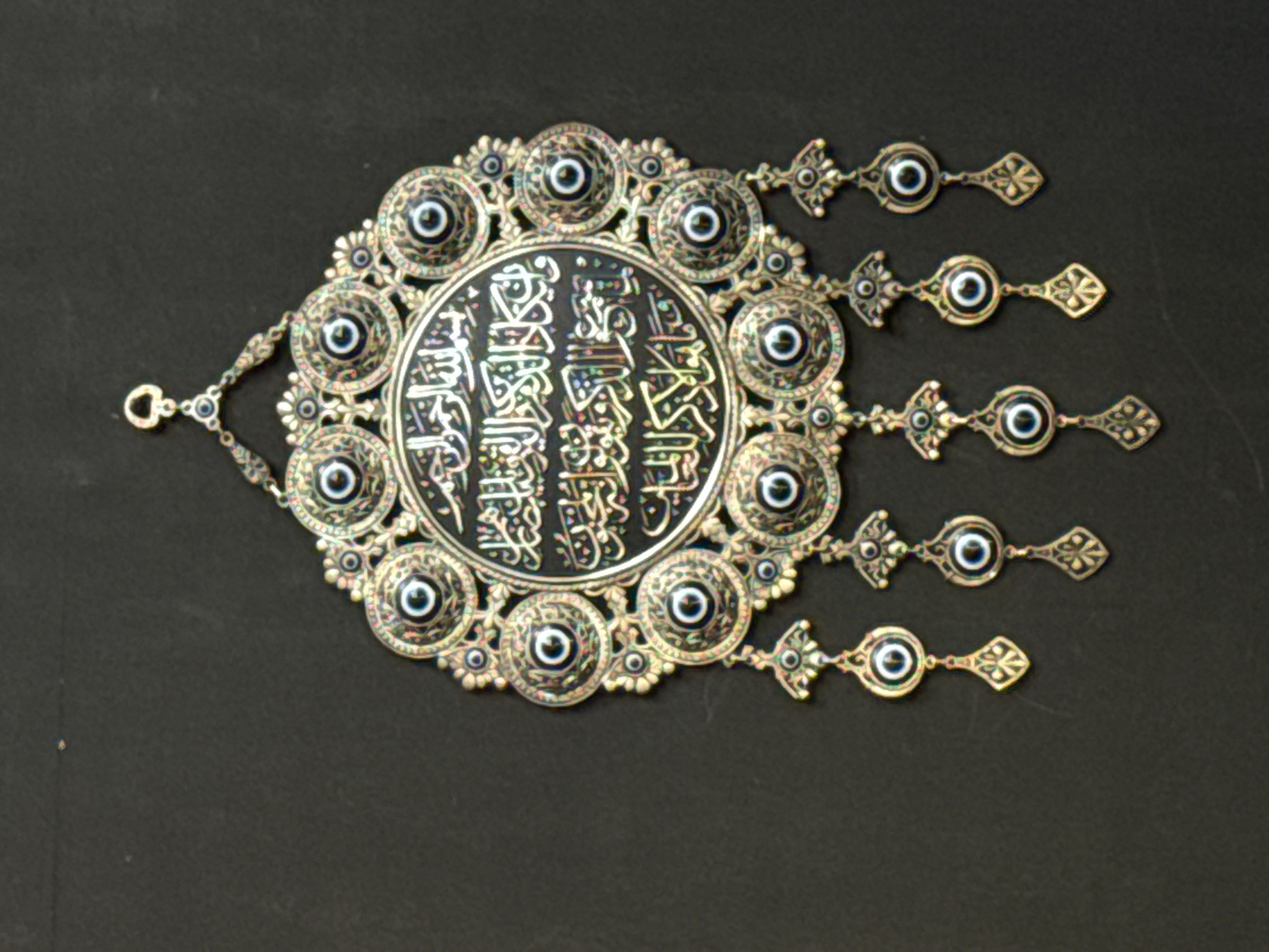 An ornate, metallic decorative wall hanging is prominently displayed against a plain, dark, solid background. The central feature is a large, circular medallion with a dark, possibly black, surface intricately inlaid with Arabic calligraphy in iridescent white or mother-of-pearl. The calligraphy initiates with "Bismillah al-Rahman al-Rahim" and continues with additional Quranic text. Surrounding this central medallion are multiple smaller, circular settings, crafted from an antique gold or brass-toned metal, each containing a concentric blue and white "evil eye" bead. The overall arrangement of these elements forms a larger, elaborate, inverted teardrop or shield-shaped framework, adorned with additional small metallic floral and geometric motifs. Five decorative metallic pendants, each mirroring the design with an "evil eye" bead set within a metallic frame and terminating in a stylized drop, suspend from the lower edge of the main structure. A single, small metallic loop is situated at the top, indicating the object is designed for hanging. The item appears to be in excellent condition, with no visible damage or significant wear. The background is uniformly dark, offering high contrast to the object's details. The lighting is even and diffused. The location is Unknown, Unknown.