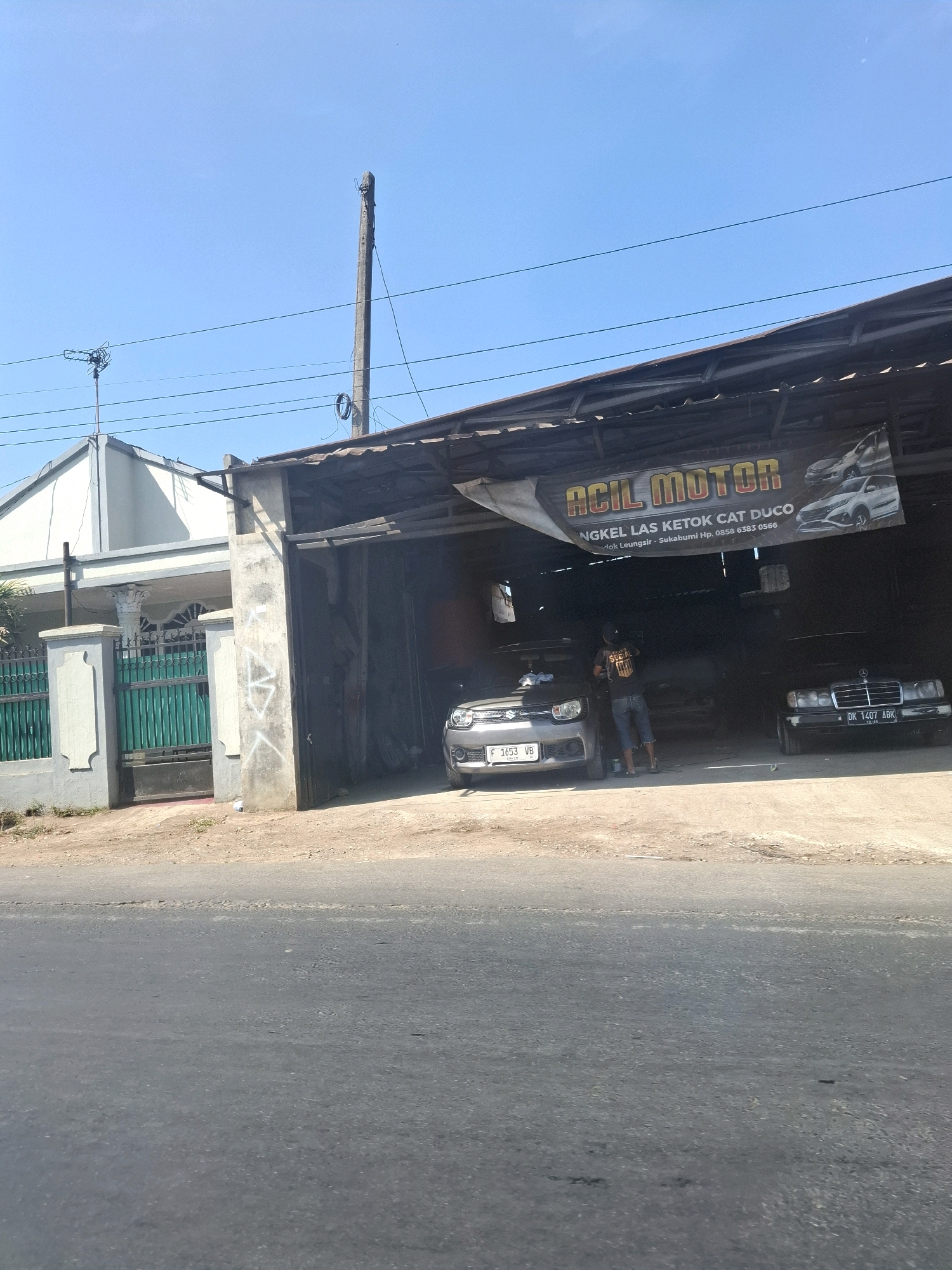 In Lembursawah, Indonesia, a street-level, eye-level shot captures an open-air auto repair shop under a corrugated metal roof. A large banner above the shop entrance reads "ACIL MOTOR" in bold yellow letters with a smaller subtitle indicating services like "LAS KETOK CAT DUCO," and contact information. Two cars are visible: a silver hatchback on the left, and a dark Mercedes-Benz sedan on the right. A person wearing a black t-shirt with "SCARED" printed on the back and blue denim shorts stands between the cars, appearing to be working. The ground is a mix of dirt and asphalt. Behind the shop, a house with a white and gray facade is partially visible, separated from the street by a green metal fence and a decorative concrete pillar. The sky is clear and blue with a single utility pole and power lines stretching across the frame. The image is taken on a sunny day.