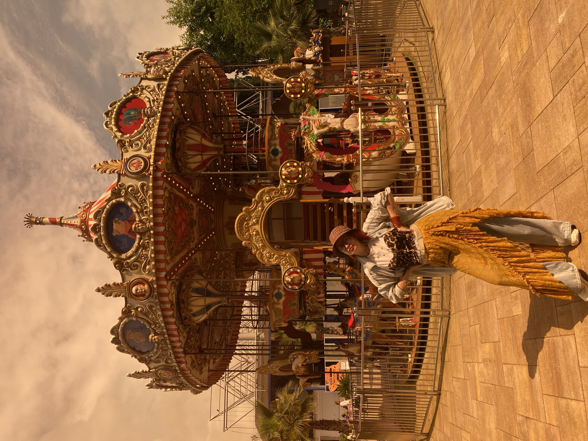 A person wearing a hat, denim jacket, a leopard-print corset-like top, and a long yellow skirt with blue denim jeans underneath stands in front of a carousel. The carousel is intricately decorated with gold trim, painted figures, and lights. It's surrounded by a metal fence. Behind the carousel, there are trees and other structures. The ground is paved with tan-colored bricks. The lighting suggests it is daytime, and the sky is visible in the upper part of the frame. This picture has been taken in Sochi, Russia.