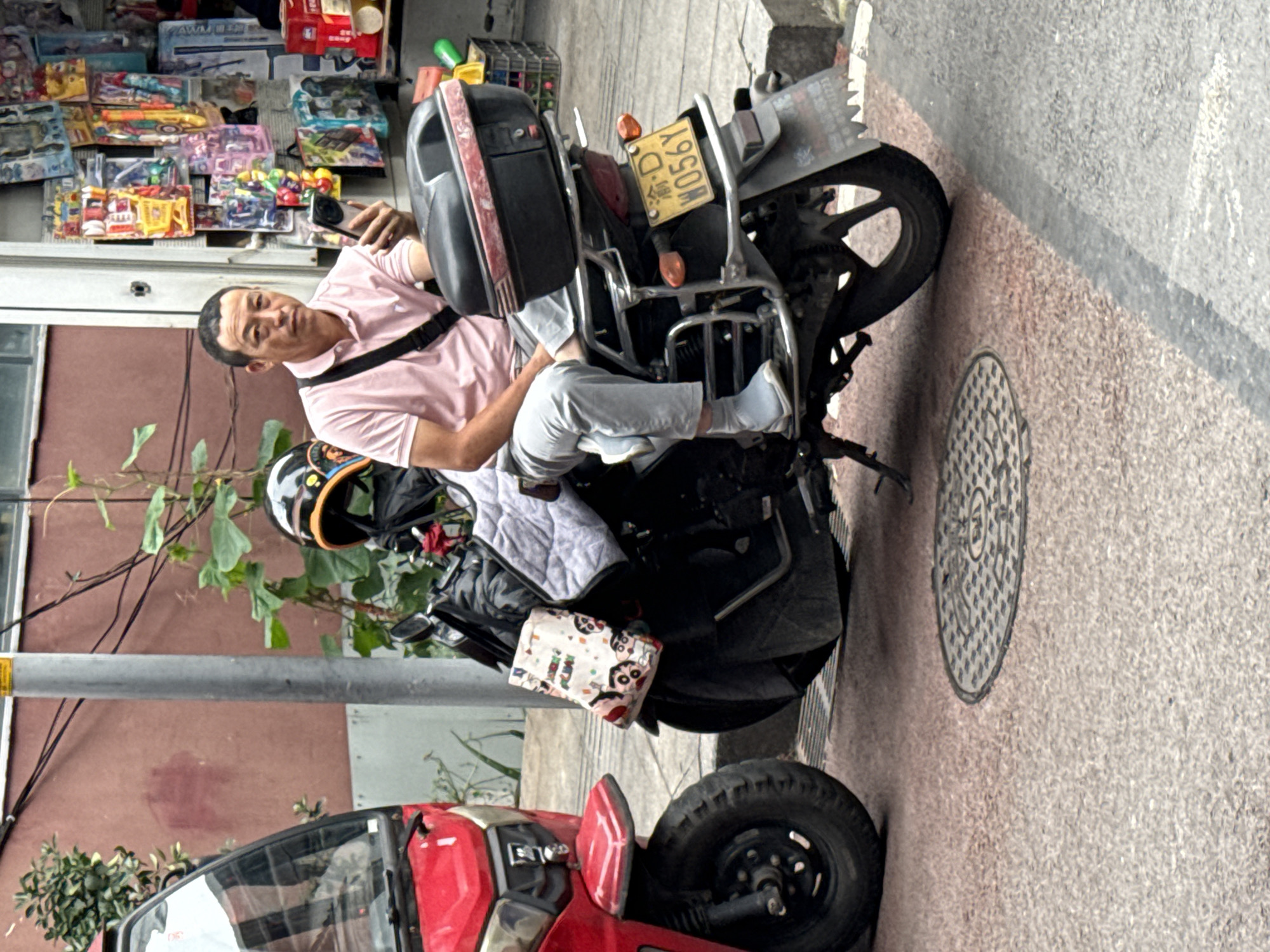 A man is straddling a motorcycle with a yellow license plate, partially parked on a sidewalk. The license plate reads "渝D W056Y". He is wearing a pink polo shirt, light-colored pants, white socks, and a black cross-body bag. He is holding a phone and looking at the camera. The motorcycle is black and has a large black box on the back. Behind the motorcycle is a storefront with shelves of toys. To the left of the motorcycle is a red three-wheeled vehicle. The scene is in the city of Jiangjin, China. A manhole cover is visible on the sidewalk. A small tree or bush is to the left of the motorcycle.
