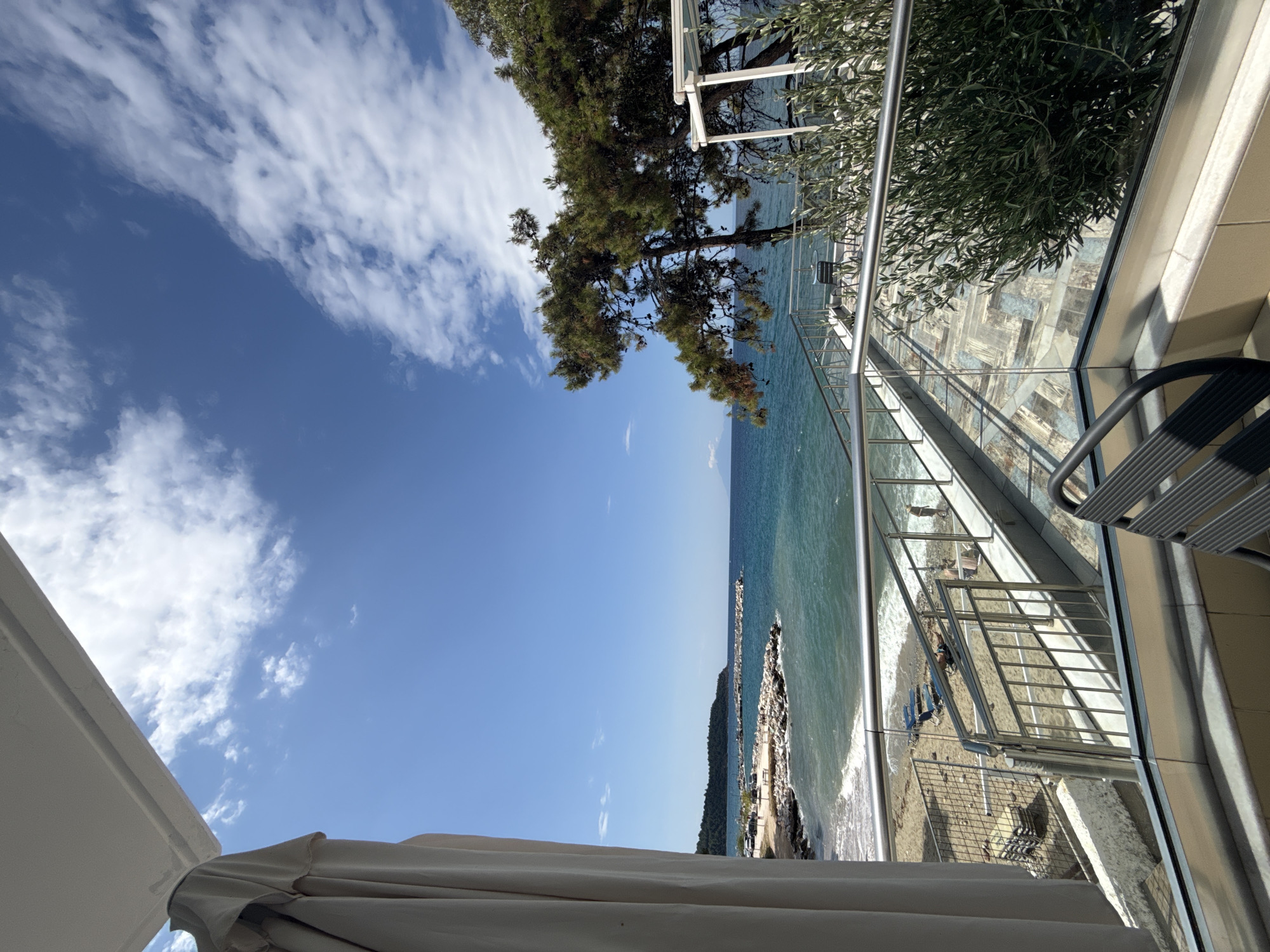 This is a photograph of a scenic view from a balcony overlooking a beach and the sea. The sky is a clear blue with scattered white clouds. On the left side of the image, a folded beige awning or canvas is visible. The balcony has a glass railing and a tiled floor.
In the middle ground, there is a sandy beach with several lounge chairs, some stacked and some in use. People can be seen on the beach, including one person standing near the water. Waves are gently lapping onto the shore. Beyond the beach, a rocky jetty extends into the turquoise sea, with several boats docked behind it. The coastline on the left is lined with trees and hills, suggesting a natural landscape.
On the right side of the image, a large evergreen tree with a spreading canopy is prominent, its branches extending over the balcony. Behind the tree, a white structure, possibly a pergola or a part of a building, is visible. In the foreground, a black chair with a slatted seat and back is partially visible, facing away from the camera. The overall scene depicts a tranquil coastal environment, likely a tourist destination. The location is Potos, Greece.