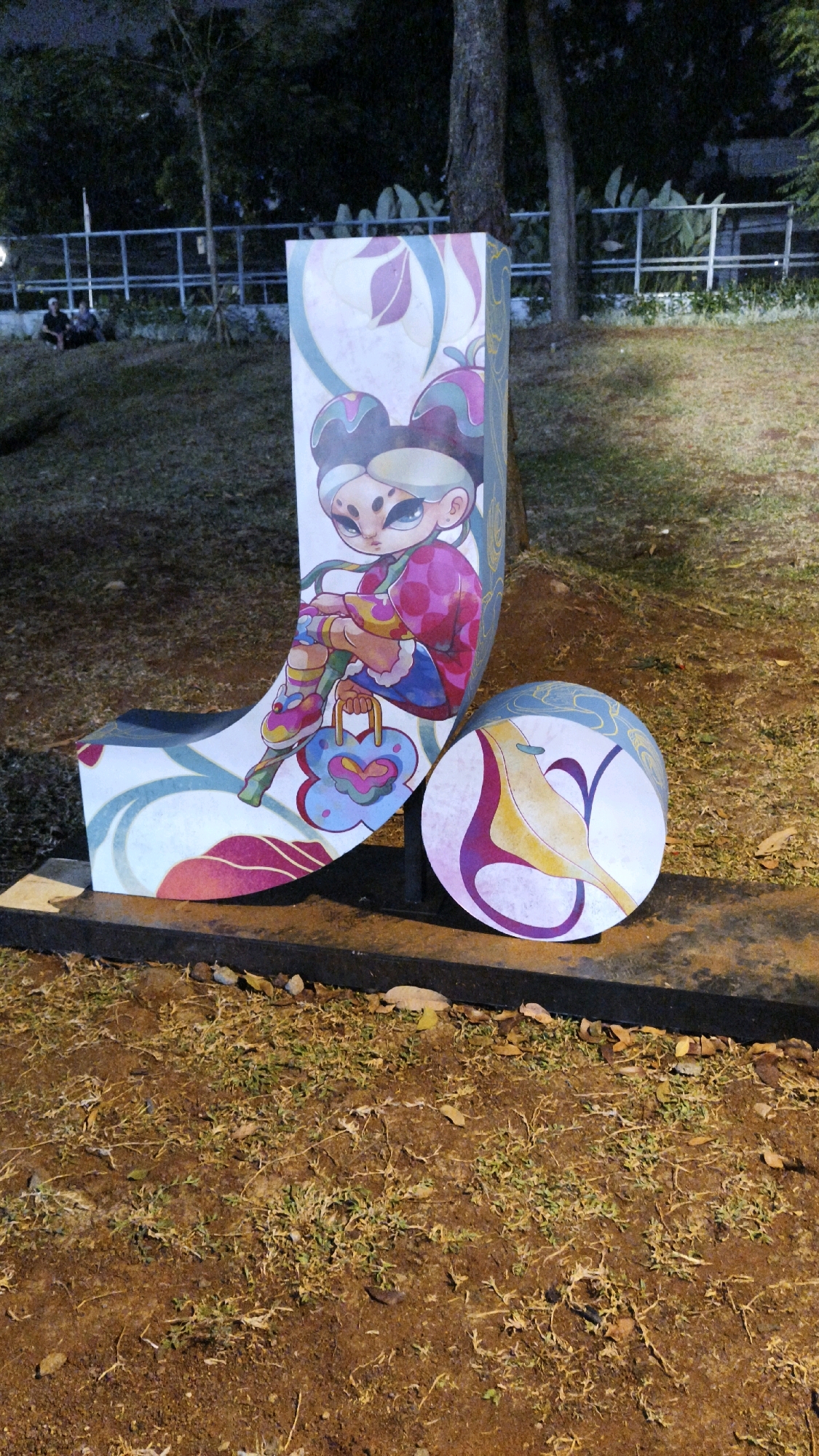 The image shows a street art installation in Taman Suropati, South Jakarta, Indonesia. The installation features a curved, bench-like sculpture with colorful artwork of a stylized character. Next to it, a circular disc also displays similar artistic elements. The sculpture and disc are placed on a concrete base, surrounded by dry soil and sparse vegetation. In the background, a metal fence separates the park area from trees and foliage. A few individuals are seated on the grass in the distance, under the shade of trees. The lighting suggests it is evening or night, with artificial lights illuminating the artwork and surrounding area.