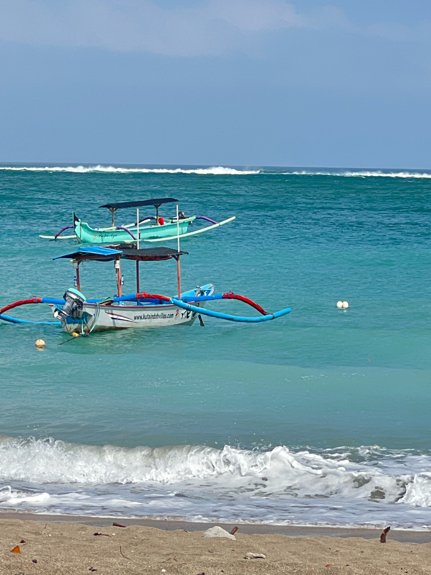 The image depicts the ocean in Kuta, Indonesia, with a sandy beach in the foreground. Two traditional Balinese fishing boats, known as jukungs, are anchored in the turquoise water. The boat in the foreground is white with blue and red outriggers, and a small outboard motor is attached to its stern. It has a canopy supported by wooden poles, offering shade. The second boat, positioned further back and slightly to the left, is a lighter shade of green and also features a canopy. In the background, white waves are breaking, indicating a moderate surf. Two white buoys are visible floating in the water between the boats. The sky is clear and blue, with a hint of haze near the horizon. The scene conveys a peaceful, tropical atmosphere typical of a beach in Kuta.