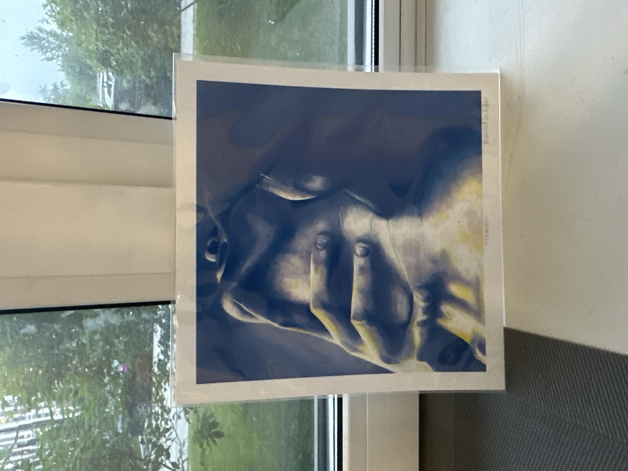 A close-up shot shows a framed artwork propped against a window frame. The artwork appears to be a print of a partial human figure in shades of blue, gray, and yellow, with hands covering the face. The artist's name, "Rachel Leifer," is written along the right edge of the print. The word "THRUST" is vertically displayed beneath the artist's name. Outside the window, lush green trees and foliage are visible. The artwork is in a clear plastic sleeve. The location is not possible to determine based on the provided image.