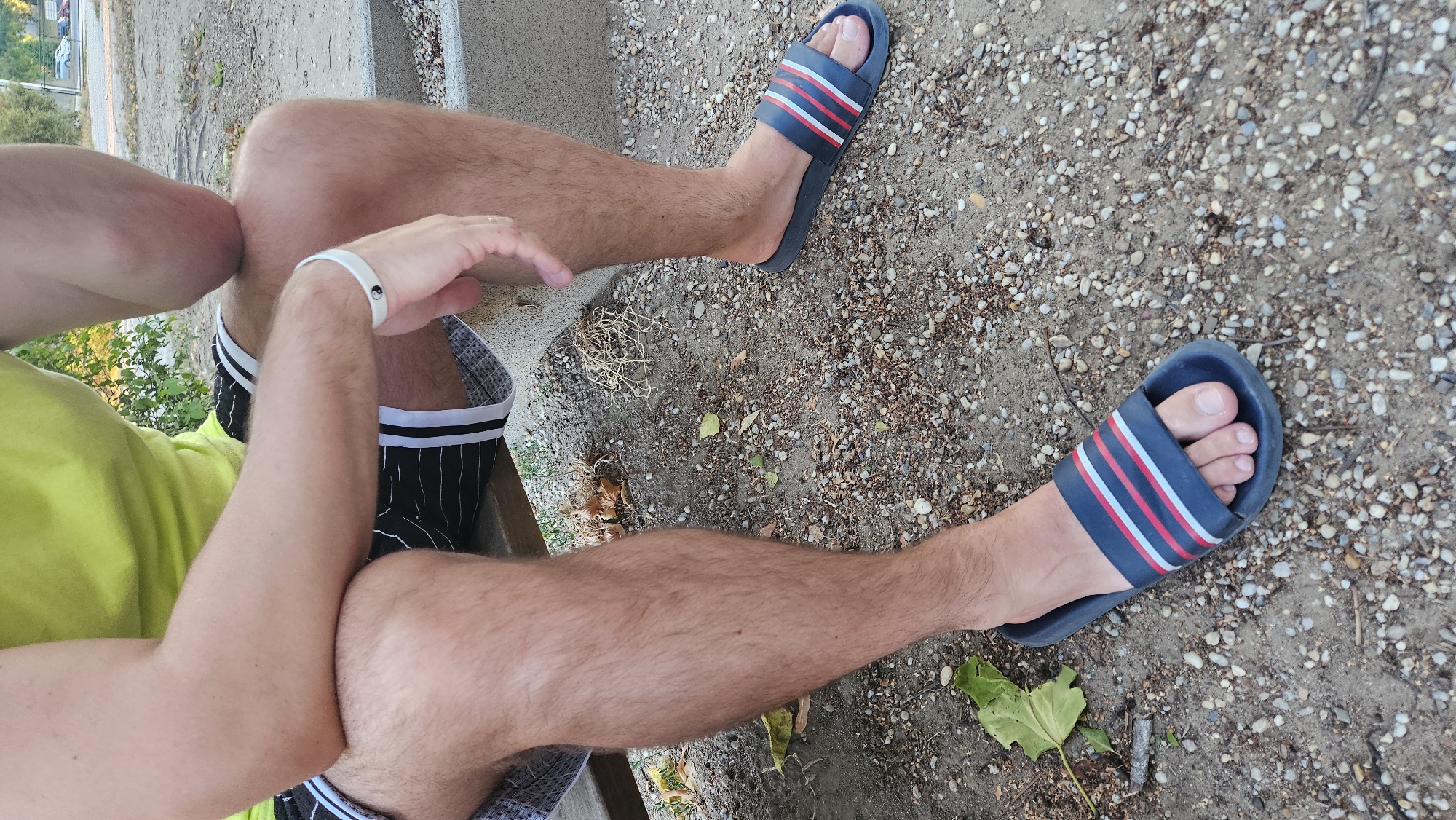 A person is sitting on a concrete bench outdoors. They are wearing a yellow t-shirt, black shorts with white stripes, and blue slide sandals with red and white stripes. The person's legs are exposed, showing short, light-brown hair. They have a white bracelet with a black yin-yang symbol on their left wrist. The ground around the bench is covered with small pebbles and scattered dry leaves. In the background, there are some green bushes and a street with parked cars and buildings visible. The scene suggests a casual outdoor setting, possibly a park or public area.