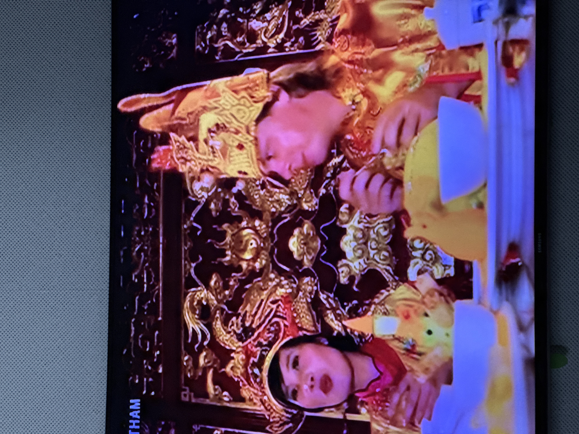 The image shows two individuals, a woman and a man, dressed in ornate golden attire and elaborate headdresses, seated at a table. The woman, on the left, has dark hair and is wearing a red and gold garment. She has bold red lipstick and is looking to her left. The man, on the right, has lighter hair and is also wearing a golden outfit and headdress. He is looking to his right. They are positioned in front of a highly decorated, dark background with intricate golden carvings, possibly depicting dragons and other mythical creatures. In the foreground, there is a table with food, including what appears to be yellow rice in bowls, and some decorative elements. The overall scene suggests a formal or ceremonial occasion, possibly a cultural or religious event. The image is captured on a screen, indicated by the texture and border around the picture. There is text visible at the top left of the image that reads "THAM". There is no indication of the location being Oskemen, Kazakhstan.