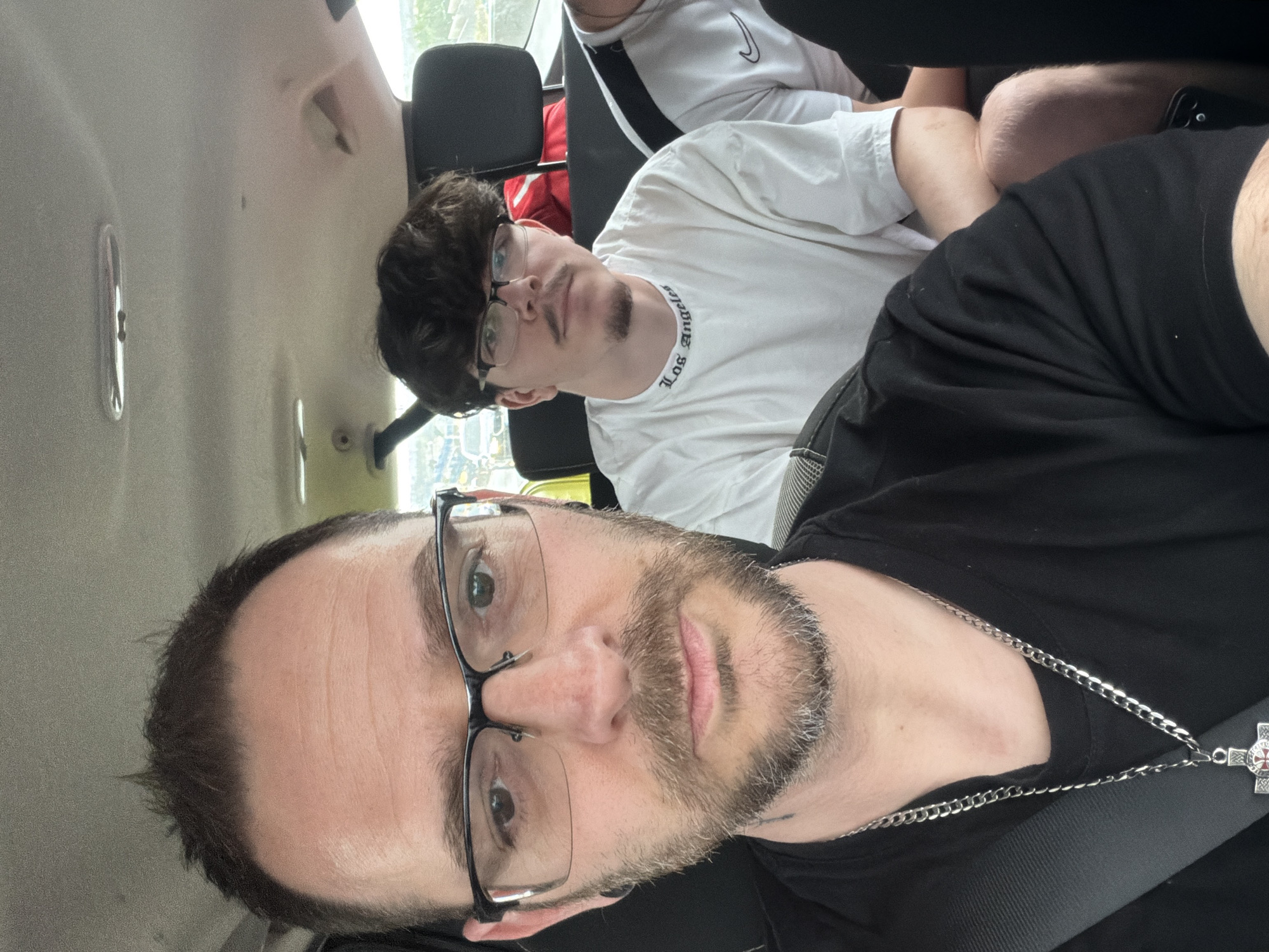 A selfie inside a vehicle shows two visible males. The foreground figure, likely the photographer, is wearing glasses and a black t-shirt, and has a silver chain with a cross pendant. Behind him sits another male, also wearing glasses and a white t-shirt with "Los Angeles" printed on the collar. Parts of other people are visible in the back seats. The interior of the car suggests an urban setting. Based on visual cues like the vehicle interior and the architecture visible through the windows, and the context provided, the photo was taken in Matosinhos, Portugal. The specific location in Matosinhos is not discernible without more visible landmarks. The action is passengers sitting inside a vehicle.