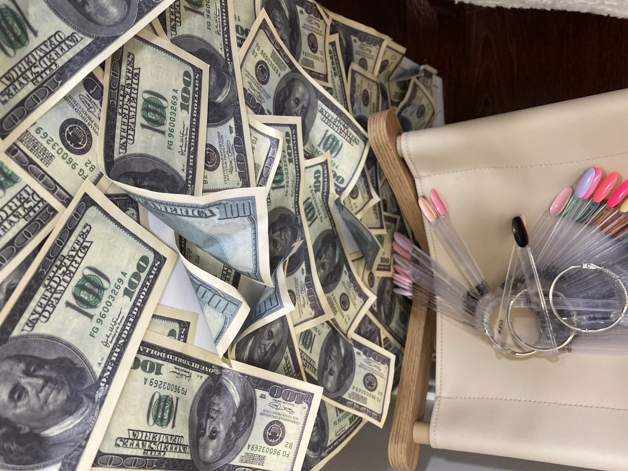 A high-angle shot shows a large pile of American one-hundred-dollar bills. Next to the pile of money is a beige leather handbag. On the handbag are several transparent plastic sticks with different colors of nail polish at the end. Silver hoop earrings lay on top of the nail polish samples. The environment is not identifiable in this image, and there is no indication of Sochi, Russia.