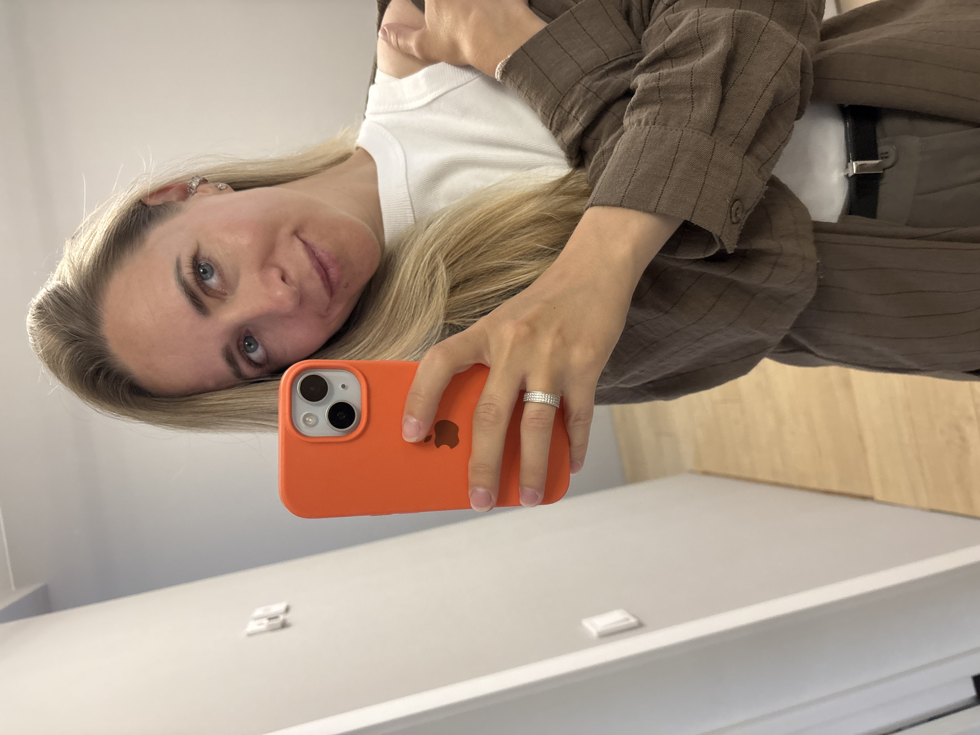A woman with blonde hair and blue eyes takes a mirror selfie in an indoor setting. She is wearing a white tank top, a brown pinstripe jacket, and grey pants with a black belt. She holds an orange iPhone in her right hand, and a silver ring with small diamonds is visible on her left ring finger. The background shows a light grey wall and a wooden floor. The image appears to have been taken in a residential apartment or hallway. There is no identifiable information to suggest the location is in Chaikovsky, Russia.