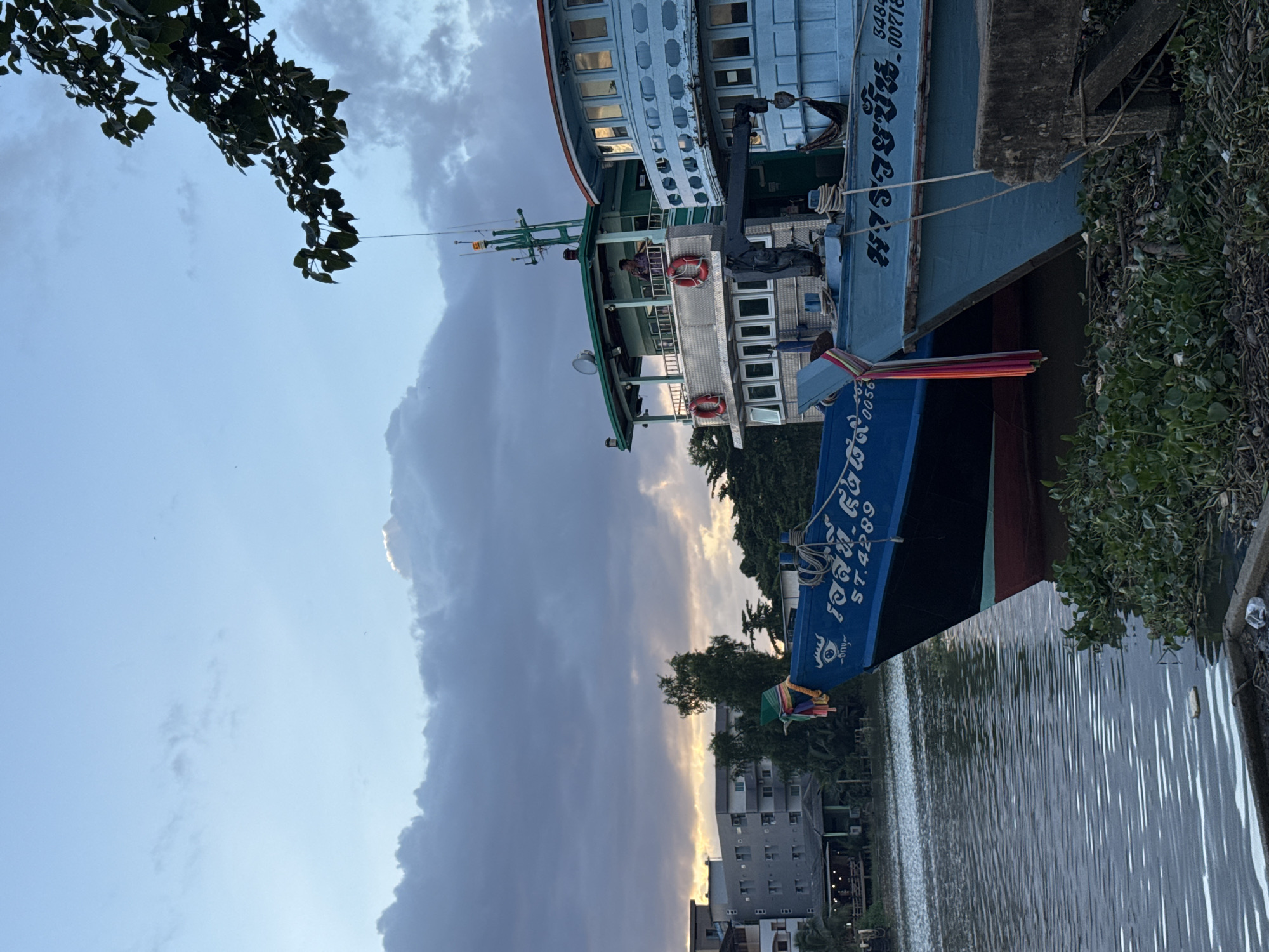 This image shows a riverfront scene in Surat Thani City Municipality, Thailand, likely at dusk. Two blue fishing boats are moored side-by-side. The boat in the foreground has Thai script and the registration number "ST.4289" visible on its hull. A man stands on the upper deck of the boat behind it, which is green and has life preservers on its railings. The sky is partly cloudy with a sliver of sunlight breaking through the clouds near the horizon. In the background, a multi-story building and trees are visible along the riverbank. The water is calm with some aquatic vegetation growing near the edge of the frame.