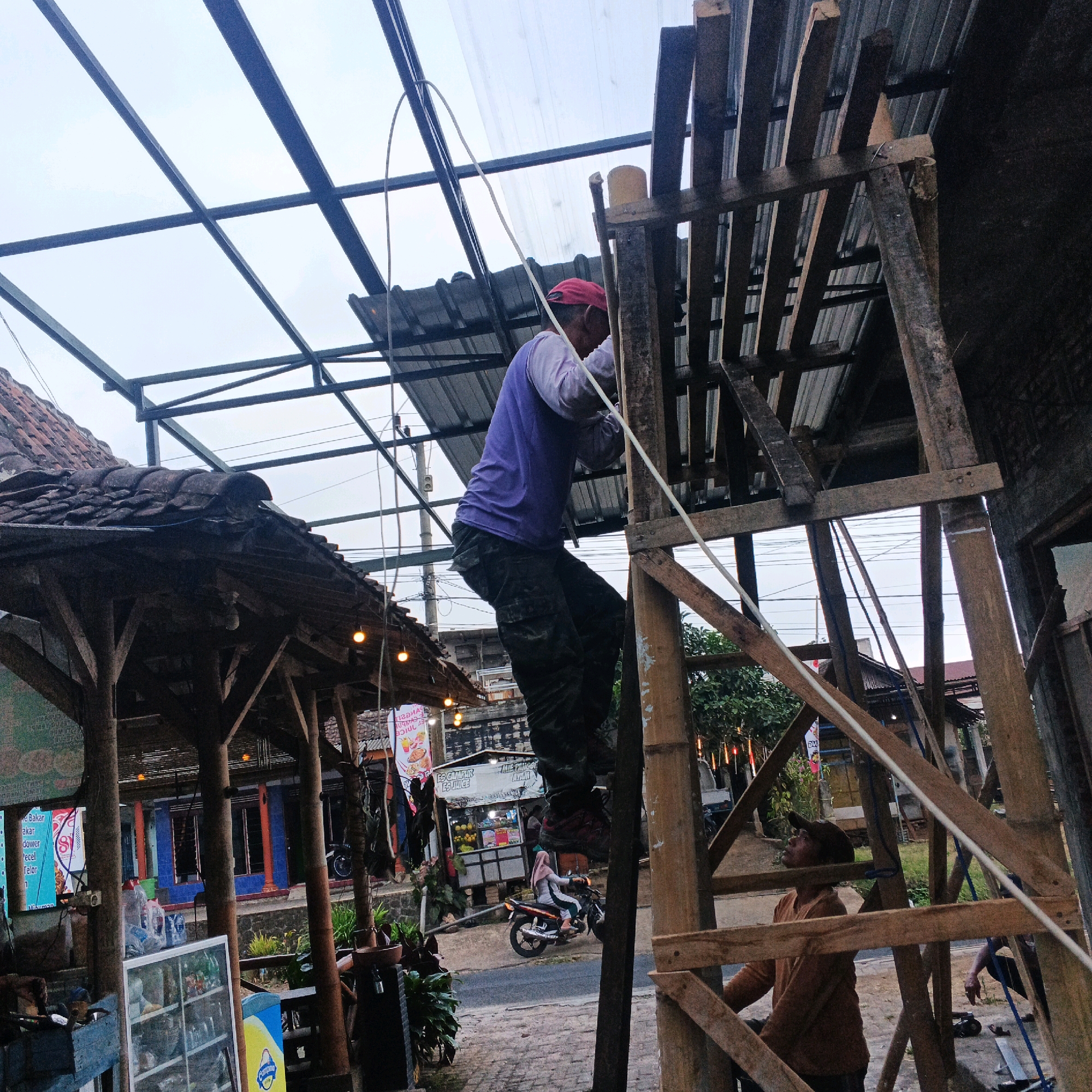 The image depicts a construction scene in Bocek, Indonesia, focused on building or repairing a roof structure. A man, wearing a purple long-sleeved shirt and camouflage pants, is positioned high on a wooden scaffolding, seemingly working on the roof frame. He is wearing a red cap and appears to be actively engaged in the construction. Another man in an orange shirt is visible lower down on the scaffolding, looking upwards.
The structure being worked on features a metal frame that will likely support a roof, with a translucent sheet of material already attached in some sections. Adjacent to the construction, a building with a tiled roof and wooden supports is visible, along with a street that has a motorcycle and people in the background. The overall environment suggests an outdoor setting, likely a market or roadside establishment, given the presence of food stalls and signs in Indonesian. The sky is overcast, indicating a cloudy day. The scene conveys a sense of ongoing labor and community infrastructure development.