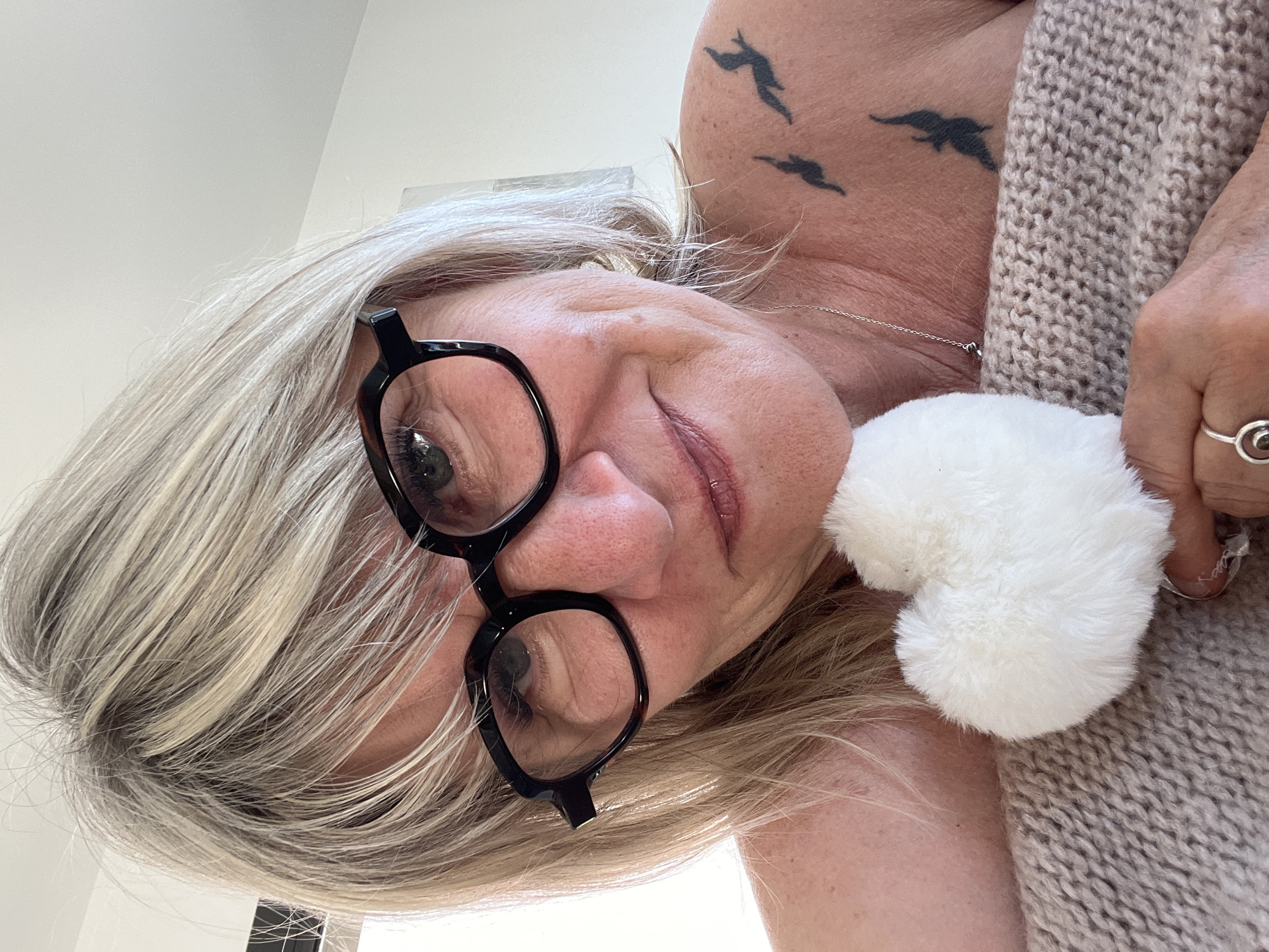 A close-up, tilted shot features a woman with short, blonde/gray hair wearing black-rimmed glasses. The subject is looking directly into the camera with a slight smile. A white, fluffy heart-shaped object is visible near her face, and she is wearing a light brown, knitted garment. On her upper chest/shoulder area, there is a tattoo depicting three birds in flight. The location is not discernable from the image. There is no way to confirm the location is Chambœuf, France.