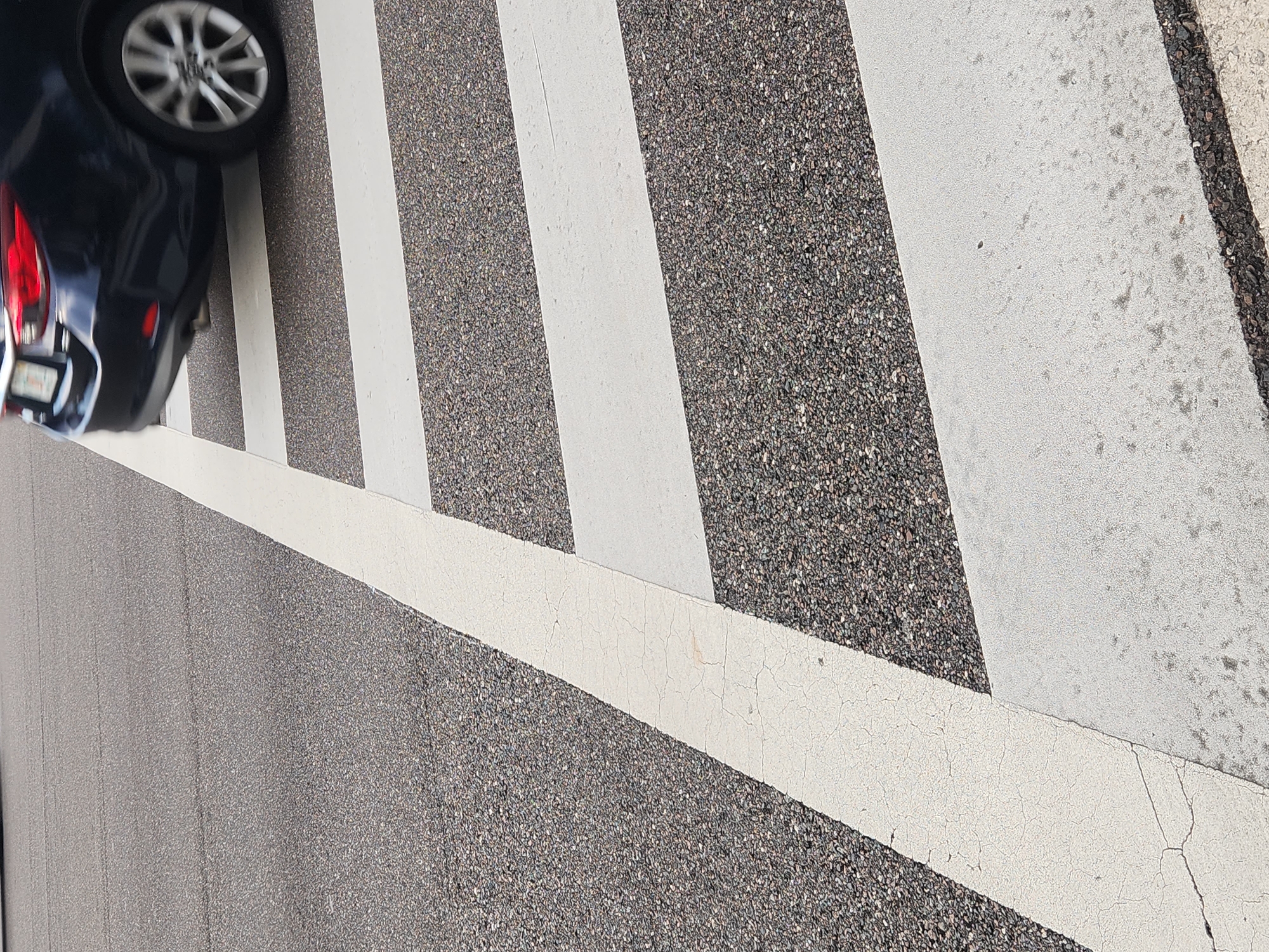 A dark-colored car is partially visible on the left side of the image, with its rear bumper and wheels showing. The car appears to be on a road or a parking lot, as indicated by the asphalt surface with white painted stripes. The stripes are typical of a crosswalk or a parking space. The image is taken from a high angle, looking down at the scene. The lighting suggests it is daytime, and the weather appears to be clear. There is no visible text in the image. The scene is static, with no discernible activity taking place beyond the presence of the car. The focus is on the textures of the asphalt and the painted lines, with the car serving as a secondary element. The overall impression is a mundane urban or suburban environment.