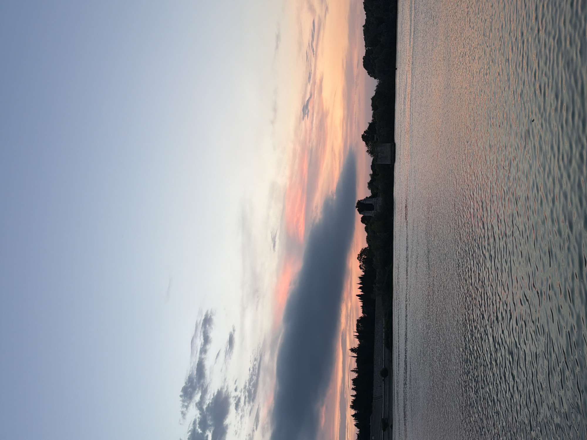The image depicts a serene sunset over the Rhône River in Arles, France. The sky is painted with soft hues of blue, pink, and orange, with scattered clouds adding texture. A dark, elongated cloud stretches across the horizon, tinged with the warm colors of the setting sun. The water of the Rhône River is calm, reflecting the colors of the sky and creating a tranquil atmosphere. Along the riverbank, a dense line of trees forms a dark silhouette against the sky. In the distance, the ruins of an old structure, possibly a tower or part of a bridge, are visible amidst the trees, adding a historical element to the landscape. The overall scene is one of natural beauty and peacefulness, characteristic of a sunset in the Provençal region.