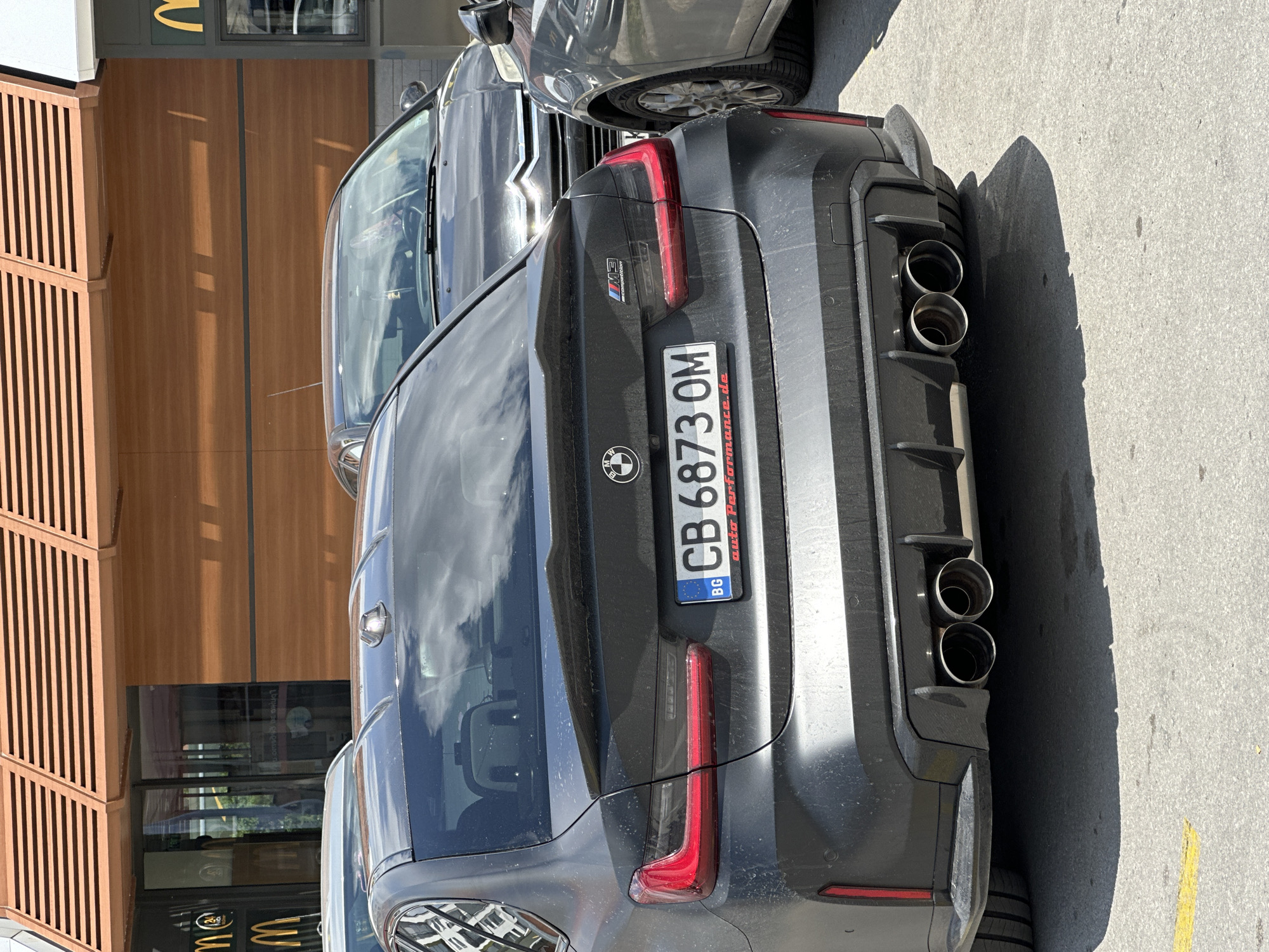A dark gray BMW M3 Competition sedan is parked in front of a McDonald's restaurant with a wooden facade in Veliko Tarnovo, Bulgaria. The car's license plate reads "CB 6873 OM". The rear of the car is visible, featuring quad exhaust pipes and a spoiler. A grey Citroën is parked behind the BMW, and another grey car is partially visible to the right. The McDonald's signage is also visible in the background. The scene appears to be a sunny day, with shadows cast on the ground.