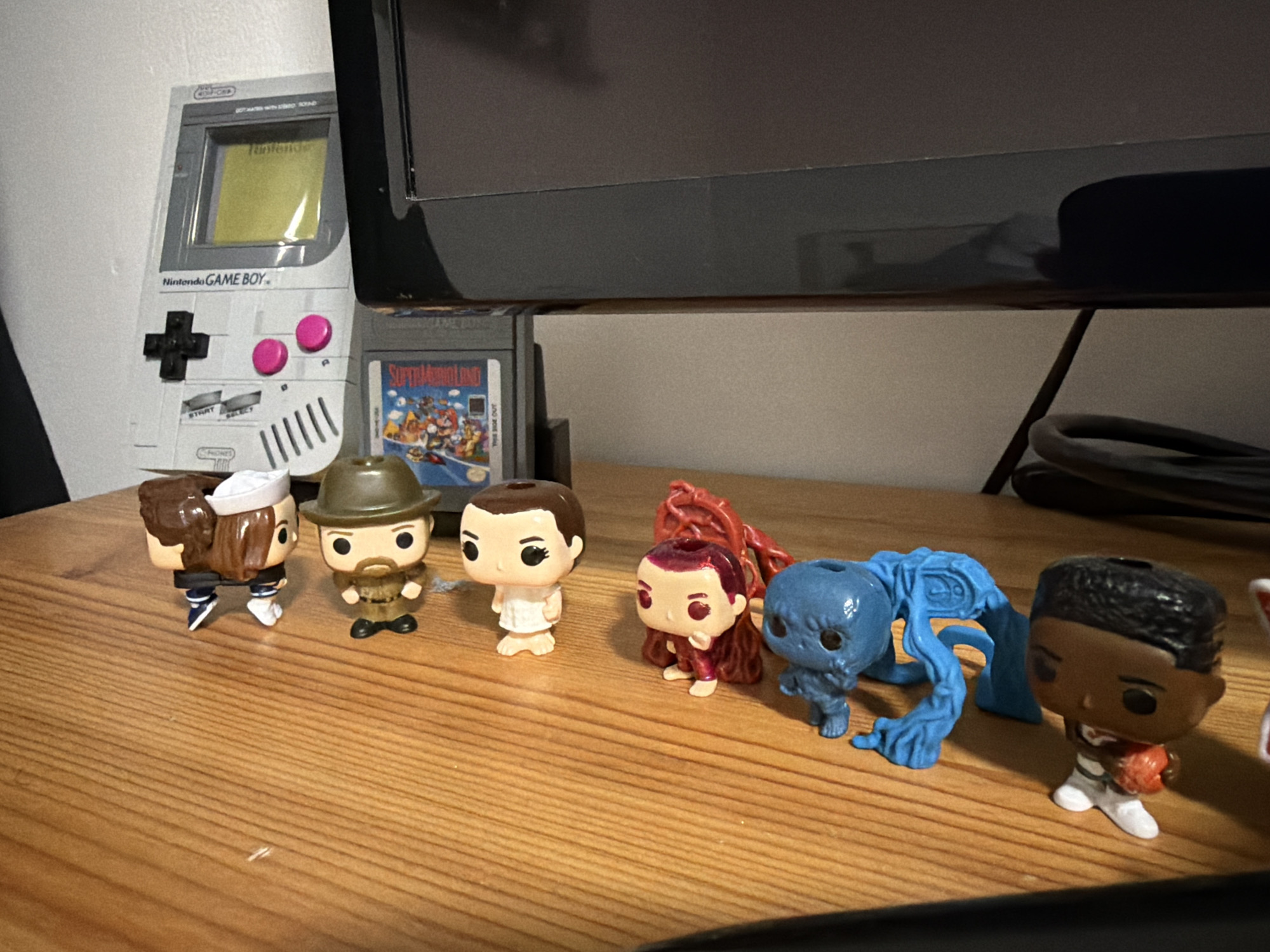 In Sankt Augustin, Germany, a collection of Stranger Things Funko Pop figures are arranged on a wooden surface. From left to right, a figure in a sailor outfit is visible, followed by one wearing a brown hat and jacket. Next is a figure in a white dress, then a figure in a red outfit with long, flowing hair. A blue, creature-like figure is positioned behind the red-haired figure, and a figure holding a basketball in white athletic wear is at the far right. Behind the figures, a grey Nintendo Game Boy and a Super Mario Land game cartridge are placed against a white wall. The scene appears to be indoors, possibly on a desk or shelf, with a dark monitor in the background.