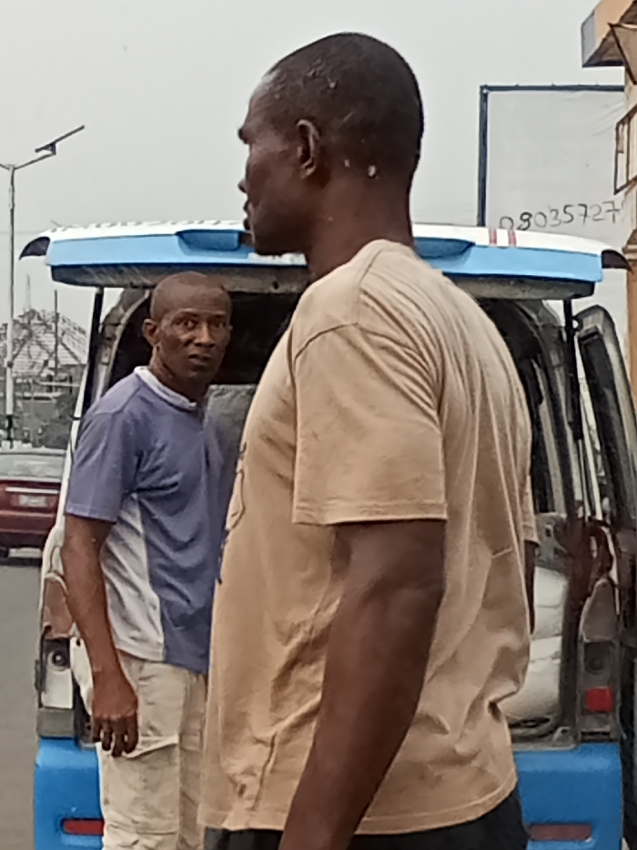 The image displays an outdoor street scene in Owerri, Nigeria, under an overcast sky. Two adult males are present near a parked commercial vehicle.
In the foreground, an adult male with dark skin and a short haircut is shown from the back-right, in profile, facing towards the right side of the frame. He wears a plain tan short-sleeved t-shirt. Directly behind him, slightly to the left, a second adult male with dark skin and a short haircut stands, looking directly at the camera with a neutral expression. He is wearing a blue and white two-toned short-sleeved shirt and light-colored trousers.
Behind the two men is a white commercial van or minibus, distinguished by a blue trim on its roof rack/spoiler area and a blue rear bumper. The rear passenger door on the vehicle's right side is open. In the background, to the left, a street light pole is visible. Further left, a portion of a reddish vehicle is present. To the right of the commercial van, a white rectangular sign or board displays a partially visible Nigerian phone number, "08035727." The ground appears to be paved.