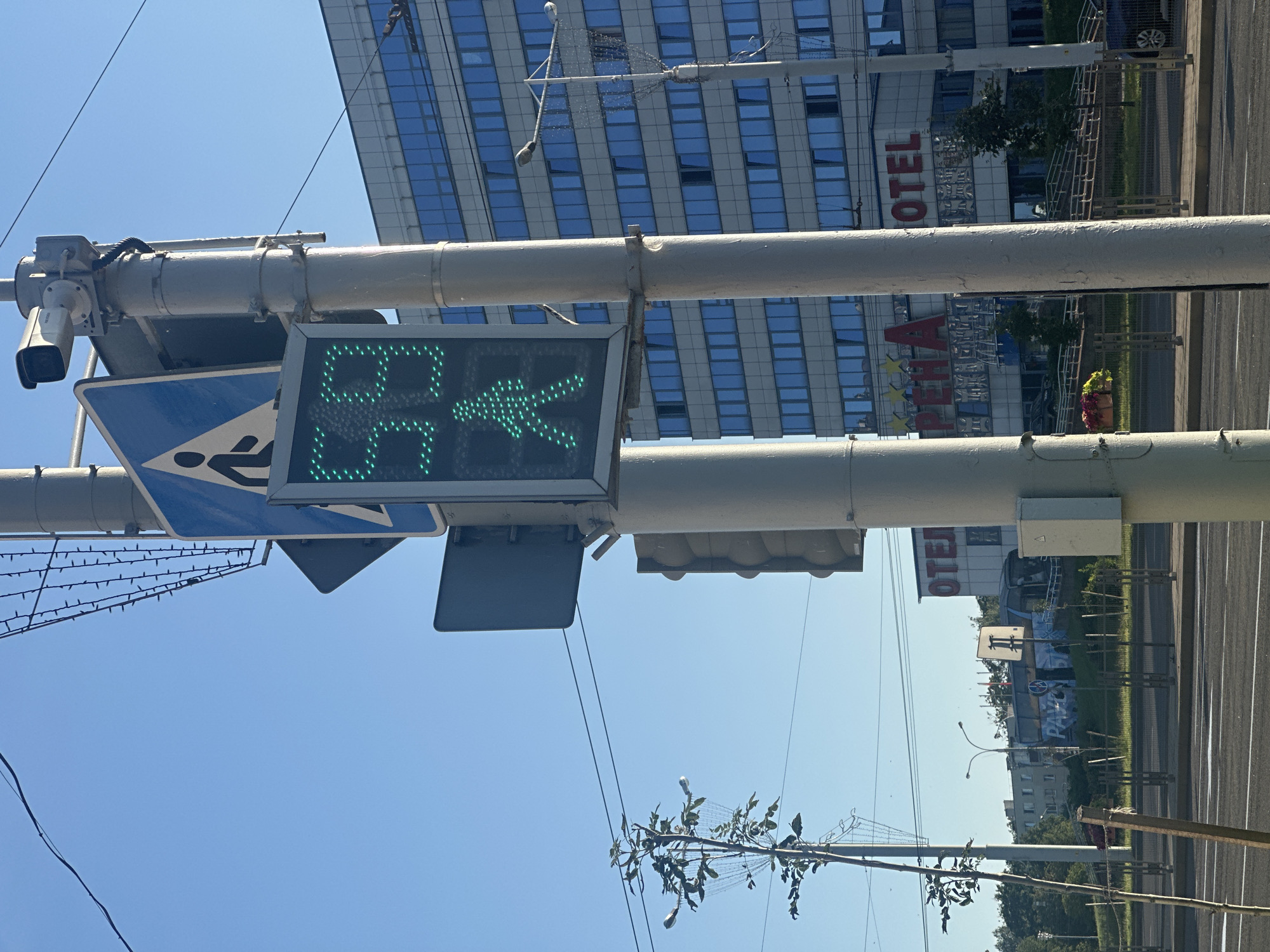 The image shows a street scene in Minsk, Belarus, on a clear day with a bright blue sky. In the foreground, a traffic light pole stands prominently. Attached to the pole is a pedestrian traffic signal displaying a green walking figure, indicating that pedestrians may cross. Above the pedestrian signal is a blue traffic sign with a white walking figure in a triangle. A surveillance camera is mounted on top of the pole. To the left of the traffic light, a thin tree with sparse leaves is visible, with decorative string lights draped around its branches.
In the background, a large modern building with many blue-tinted windows dominates the scene. The building has large red lettering on its facade that reads "ПЕРГА" and "ОTEL", suggesting it is a hotel. There are also visible signs of power lines crisscrossing the sky. Further in the background, more buildings, trees, and a street with a bus are visible. A pedestrian crossing area is implied by the presence of the pedestrian traffic signal. The overall environment suggests an urban setting, likely a busy intersection.