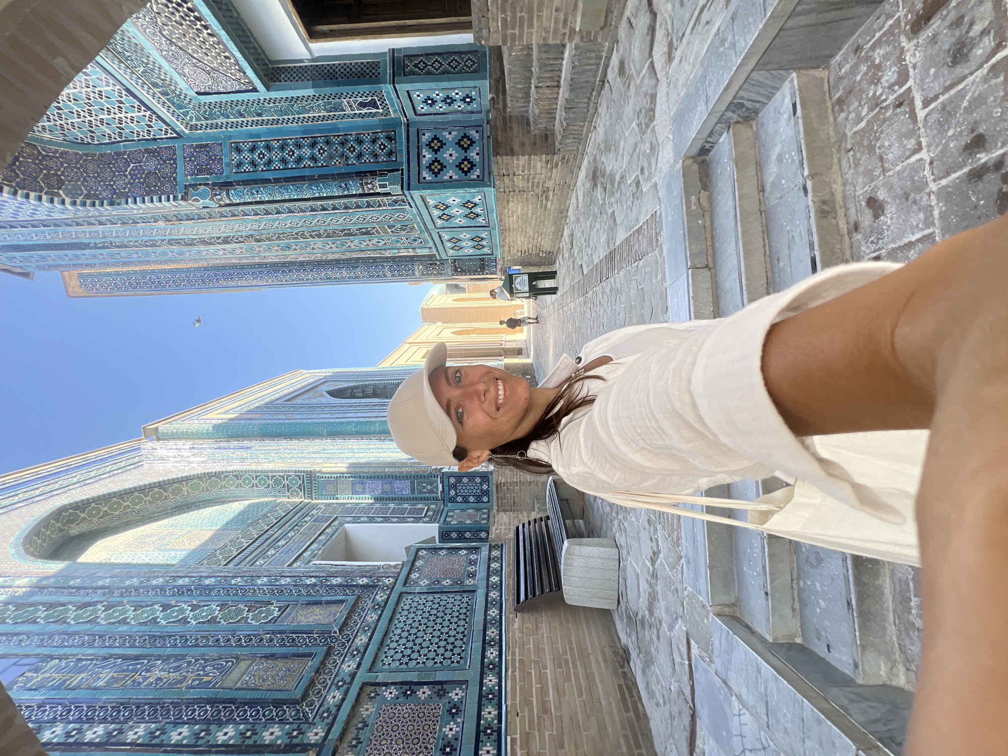 A woman wearing a cream-colored baseball cap and a white linen shirt is taking a selfie while walking through a courtyard lined with intricately tiled blue buildings. She has dark hair and is smiling at the camera. She is carrying a cream-colored tote bag. The buildings are decorated with geometric patterns and calligraphy in shades of blue, green, and white. Stone pathways and steps lead through the courtyard. In the background, other people can be seen walking. The architecture suggests this is the Shah-i-Zinda necropolis in Samarkand, Uzbekistan, known for its stunning tilework. The sky is a clear blue.