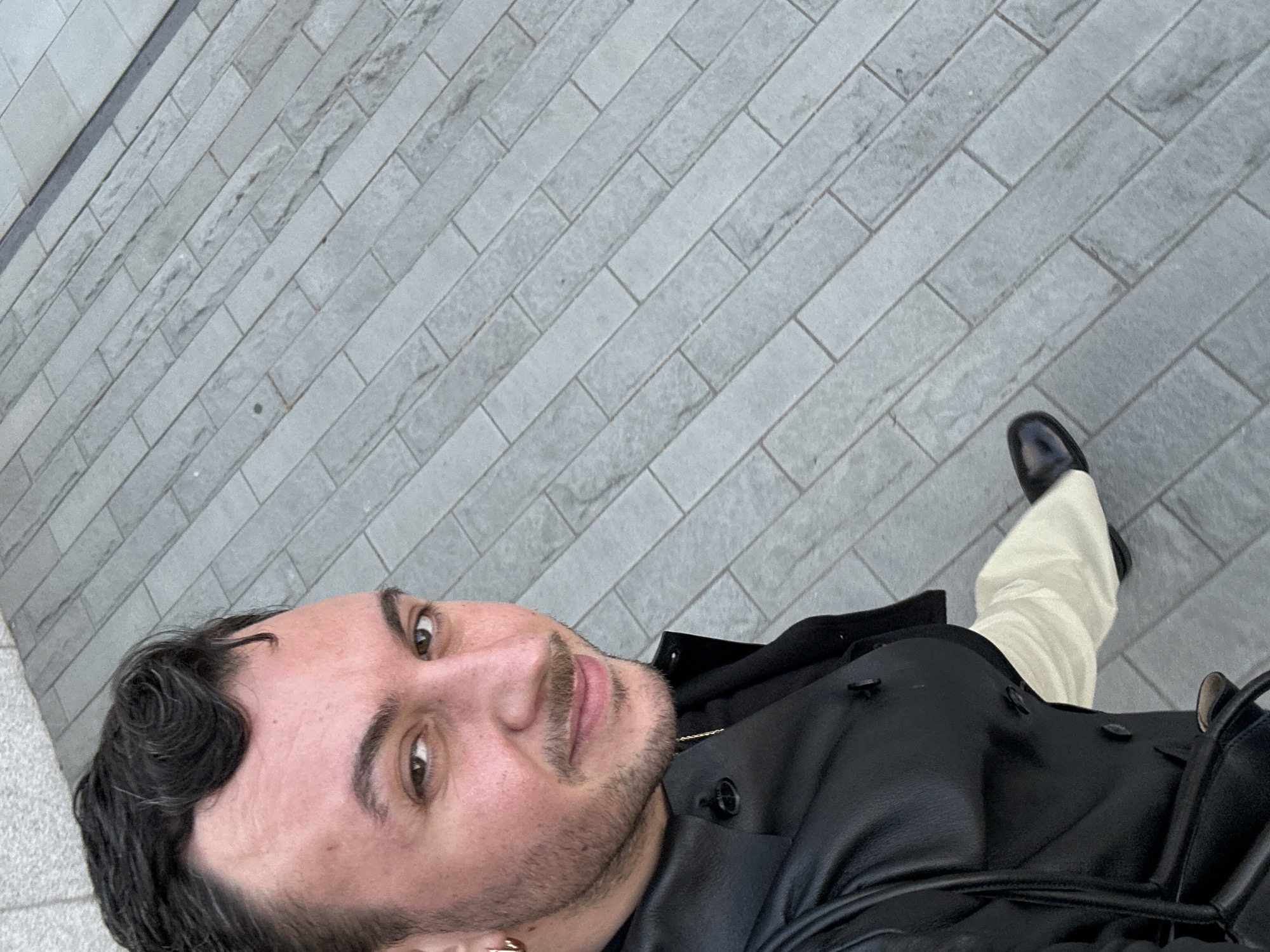 A selfie shot from above shows a man standing on a gray brick pavement. The bricks are laid out in a running bond pattern. He is wearing a black leather jacket over a black hooded sweatshirt, light-colored trousers, and a black shoe is visible. He has dark hair, stubble, and is wearing a small gold earring. The photo appears to be taken outdoors in daylight. It is not possible to determine the location of this image based on the content provided.