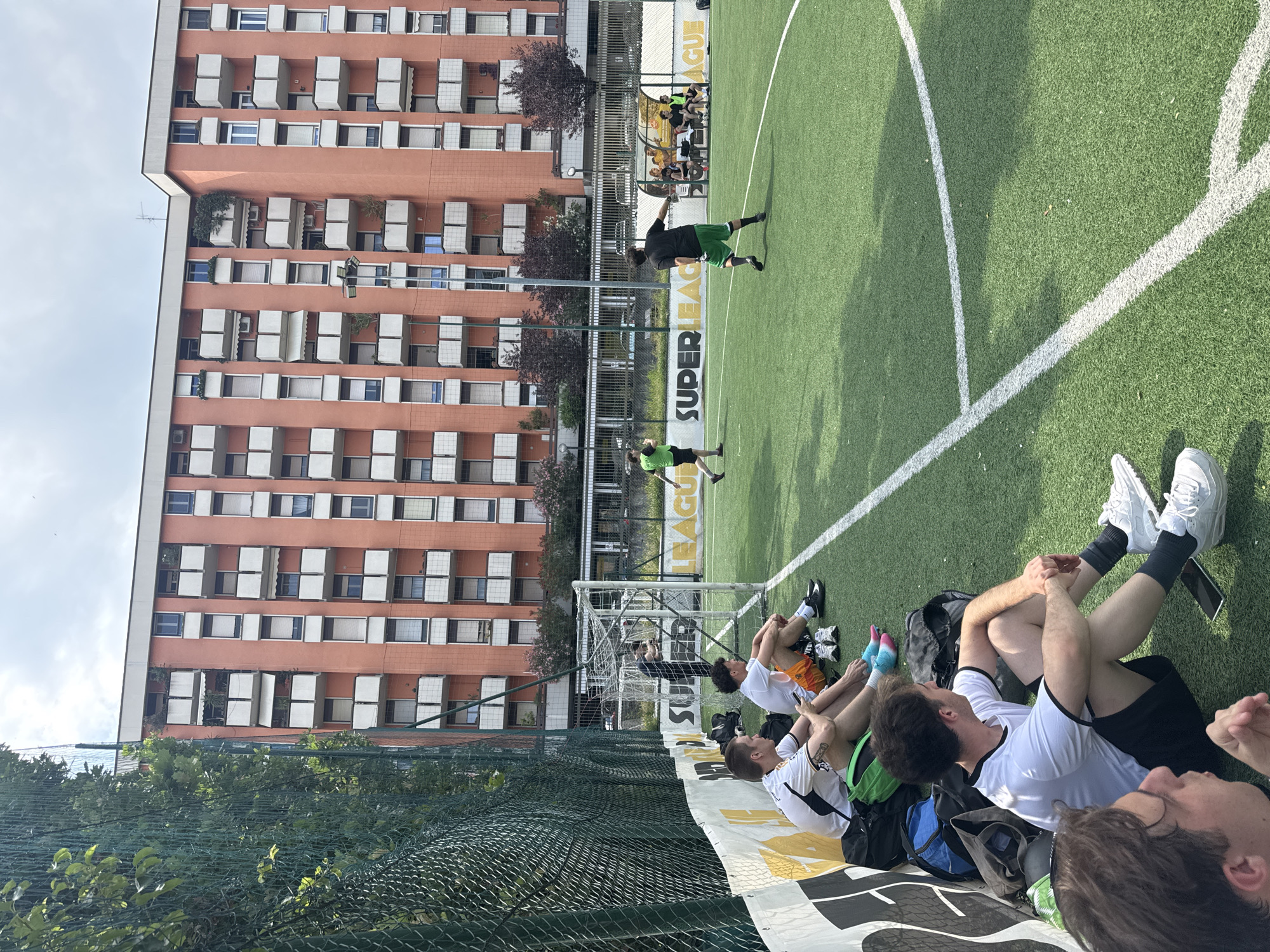 This image depicts an artificial turf football pitch located in the city of Milan, Italy, under a partly cloudy sky during daylight. In the immediate foreground on the left, three young adult males are seated on the ground along the sideline. The closest individual, with dark hair, wears a white t-shirt with black trim and black shorts, looking towards the right side of the frame. Adjacent to him, another male, similarly attired, is observing a smartphone. This individual has visible tattoos on his forearms and wears light blue and pink athletic socks; a green backpack rests nearby. A third male, wearing a white t-shirt and orange shorts, sits further back, with a pair of athletic shoes placed on the ground in front of him. Behind these seated individuals, a white metal football goal with a net is visible, and a person in dark clothing stands near it.
The football pitch itself features green artificial turf marked with white lines, including a visible section of a center circle arc and a straight sideline. On the field, two players are actively engaged. One player, seen from behind, wears a black t-shirt and green shorts, facing towards the right. Another player, in a green vest and dark shorts, walks towards the left. On the right sideline, a covered dugout structure houses approximately four individuals seated on a bench.
Along the sideline, a white banner displaying the text "SUPER LEAGUE" in black and yellow lettering is stretched. A green chain-link fence, partially obscured by green foliage, runs along the left side of the pitch. The background is dominated by a multi-story residential building with a reddish-orange facade, characterized by numerous white balconies, some featuring sun awnings or potted plants. A tall streetlight pole is positioned in front of the building, and trees with dark purple foliage are visible behind the dugout.