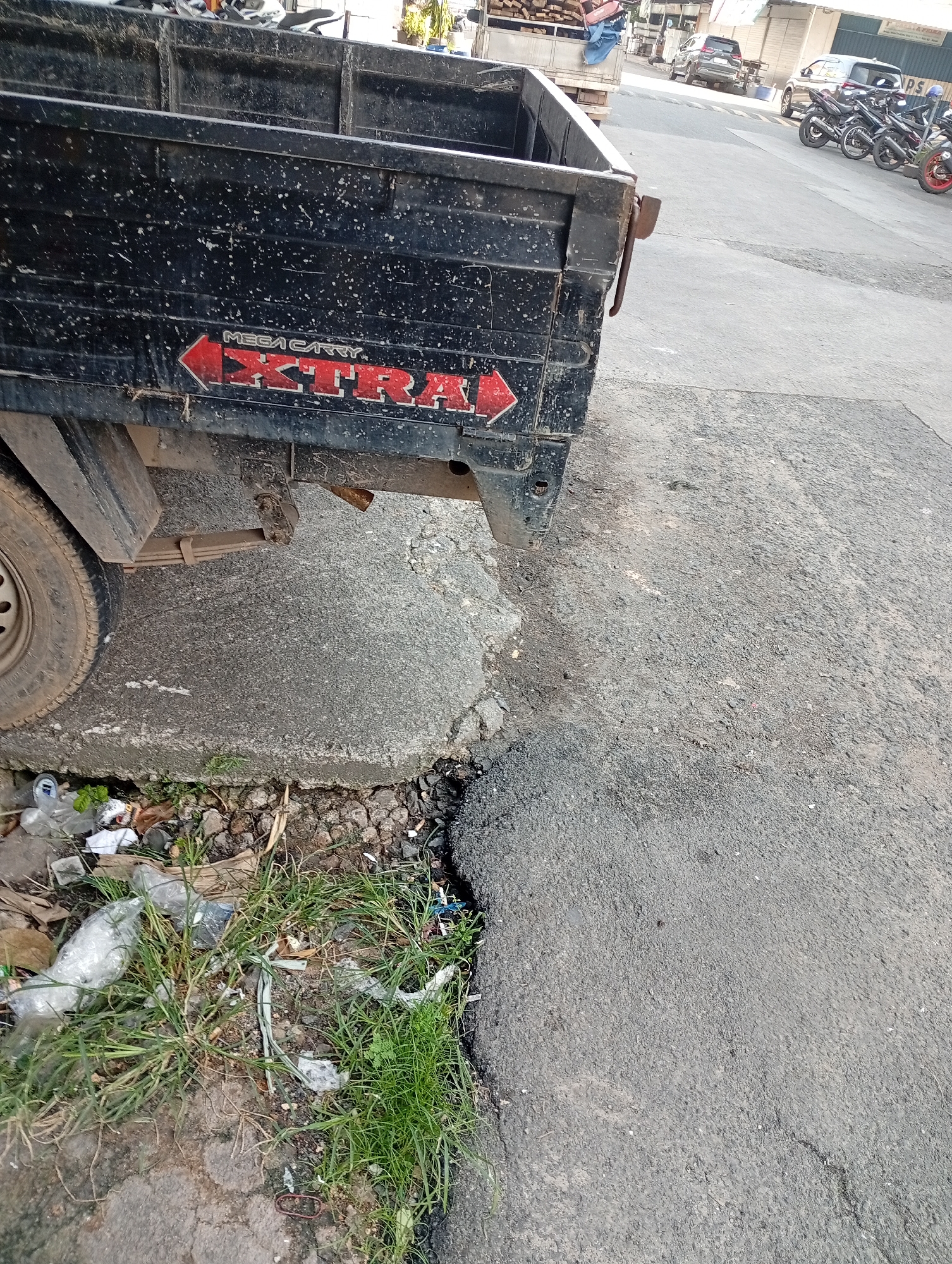In Batam, Indonesia, a black pickup truck is parked next to a broken section of asphalt road. The truck has "MEGA CARRY EXTRA" written on its side. Grass and litter, including plastic bottles and bags, are visible on the ground next to the truck. In the background, several motorcycles are parked along the street.