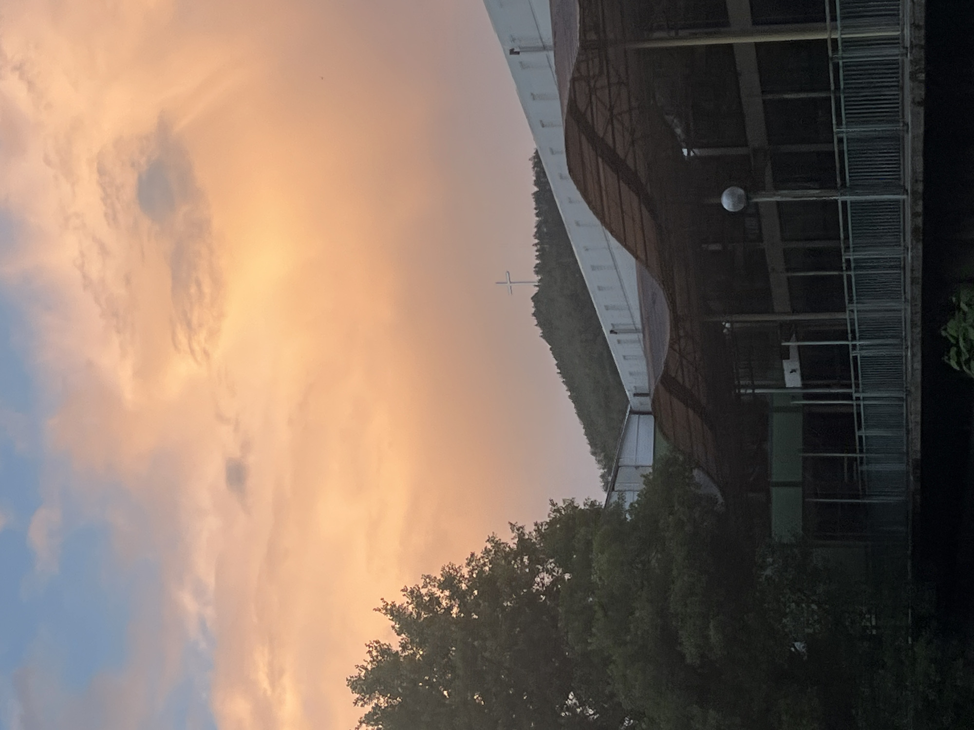 The image depicts a tranquil outdoor scene, primarily featuring a sky filled with clouds illuminated by warm light, set against a natural and urban landscape. The original image is oriented vertically, but for description, it will be considered horizontally, with the sky dominating the upper and central portions.
The sky showcases a blend of soft peach, orange, and golden hues, particularly vibrant on the left side, gradually transitioning to a lighter, more muted orange and faint blue towards the bottom right. Scattered clouds, some wispy and some more voluminous, are present, with a distinct, darker, almost silhouette-like cloud formation near the center-left. A tiny dark speck, possibly a bird or insect, is visible against the light sky in the upper right.
Below the sky, a lush, dark green canopy of trees occupies the lower foreground on the left. In the mid-ground, a densely forested hill or mountain is visible, topped by a prominent white cross, standing out against the warm sky.
To the right, a large building structure is partially visible, featuring a brown, perhaps corrugated or tiled, roof section supported by metal beams and a white facade. Below this roof, there are metal railings and what appears to be part of a multi-story building. A spherical, glowing street lamp or light fixture hangs beneath the building's roof structure. Further right and in the lower foreground, more dark green foliage is visible.
The scene suggests either sunrise or sunset, given the warm, glowing light that bathes the clouds. The overall atmosphere is serene and contemplative. No explicit activity is taking place, and there is no visible text in the image. The presence of the cross on the hill could be a significant landmark, consistent with the Blagoevgrad, Bulgaria context.