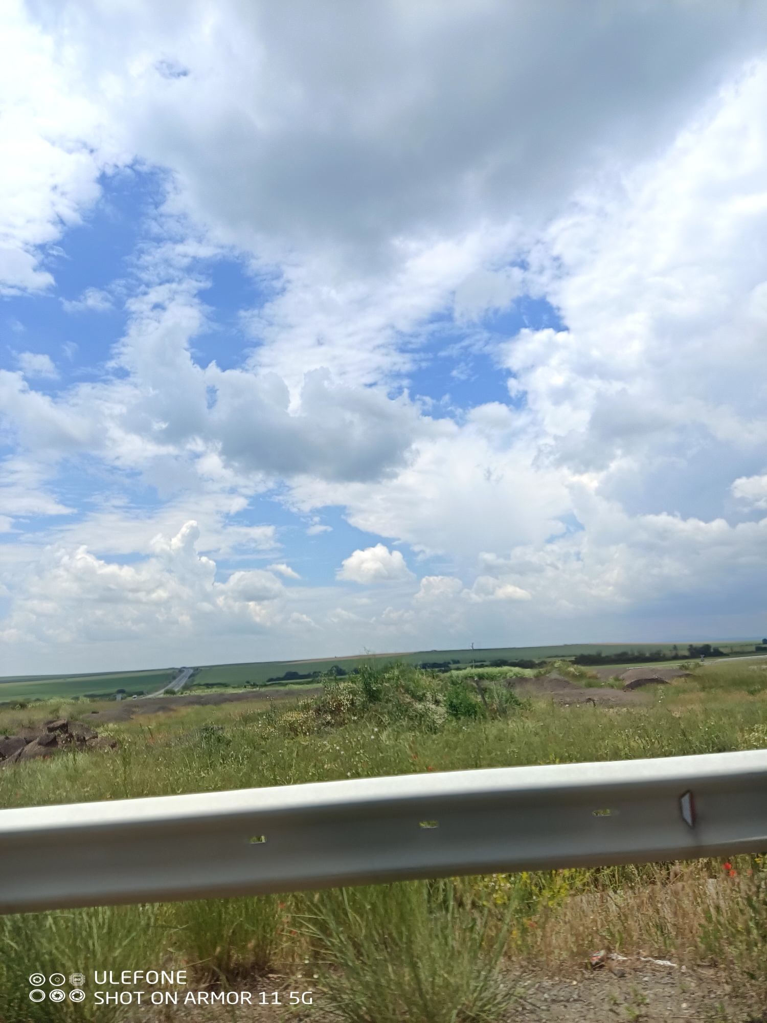 The image captures a wide-angle landscape scene on a partly cloudy day in Dragantsi, Bulgaria. A light gray metal guardrail spans the lower third of the frame, indicating the vantage point is from a roadside. Immediately in front of and below the guardrail, dry green and brown grasses with scattered small red flowers (likely poppies) are visible on a dirt and gravel shoulder. Beyond the guardrail, the terrain consists of gently rolling hills with patches of green cultivated fields and areas of disturbed brown earth, possibly from construction or excavation. A two-lane paved road or highway is visible in the mid-distance, curving through the landscape and extending towards the distant horizon. The sky is predominantly blue, filled with numerous white cumulus clouds, with some larger, denser cloud formations present. No individuals, animals, or active vehicles are discernible within the frame. The image includes a watermark: "Ulefone SHOT ON ARMOR 11 5G."