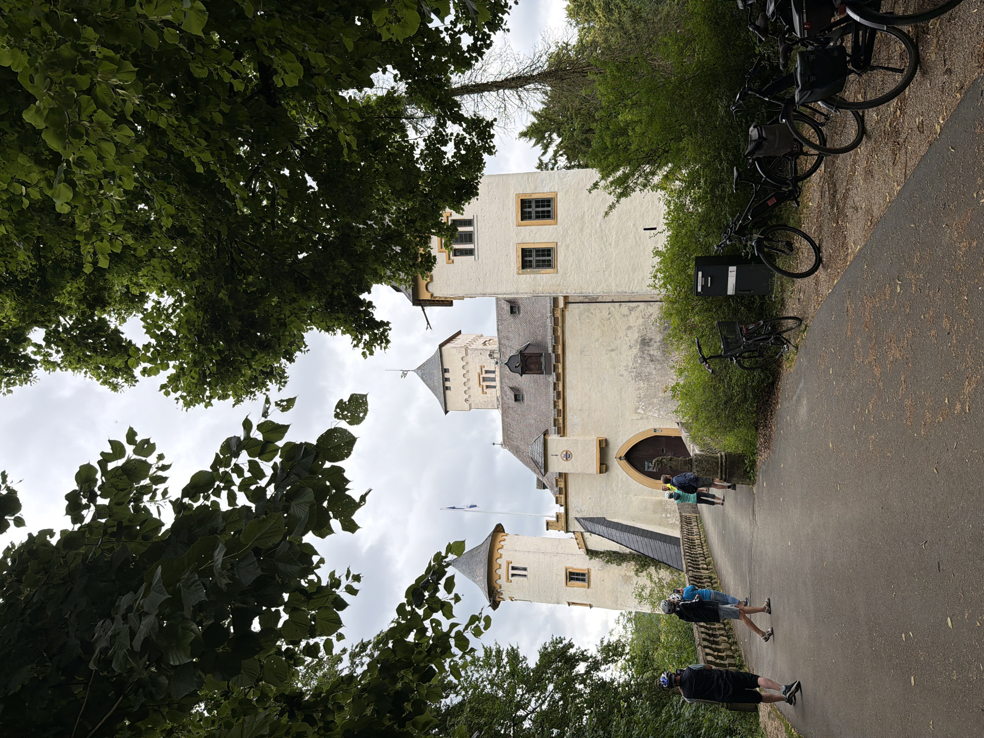 The image shows a view of a castle in Heiligenstadt in. OFr., Germany. In the foreground, a paved path leads to a stone bridge with a decorative railing. Several people, dressed in casual attire and some wearing helmets, are walking across the bridge towards the castle. To the right of the path, a row of parked bicycles is visible. The castle itself is a light-colored building with multiple towers and arched entrances. It is surrounded by lush green trees, with the leaves of some trees hanging into the frame from the top. The sky is overcast.