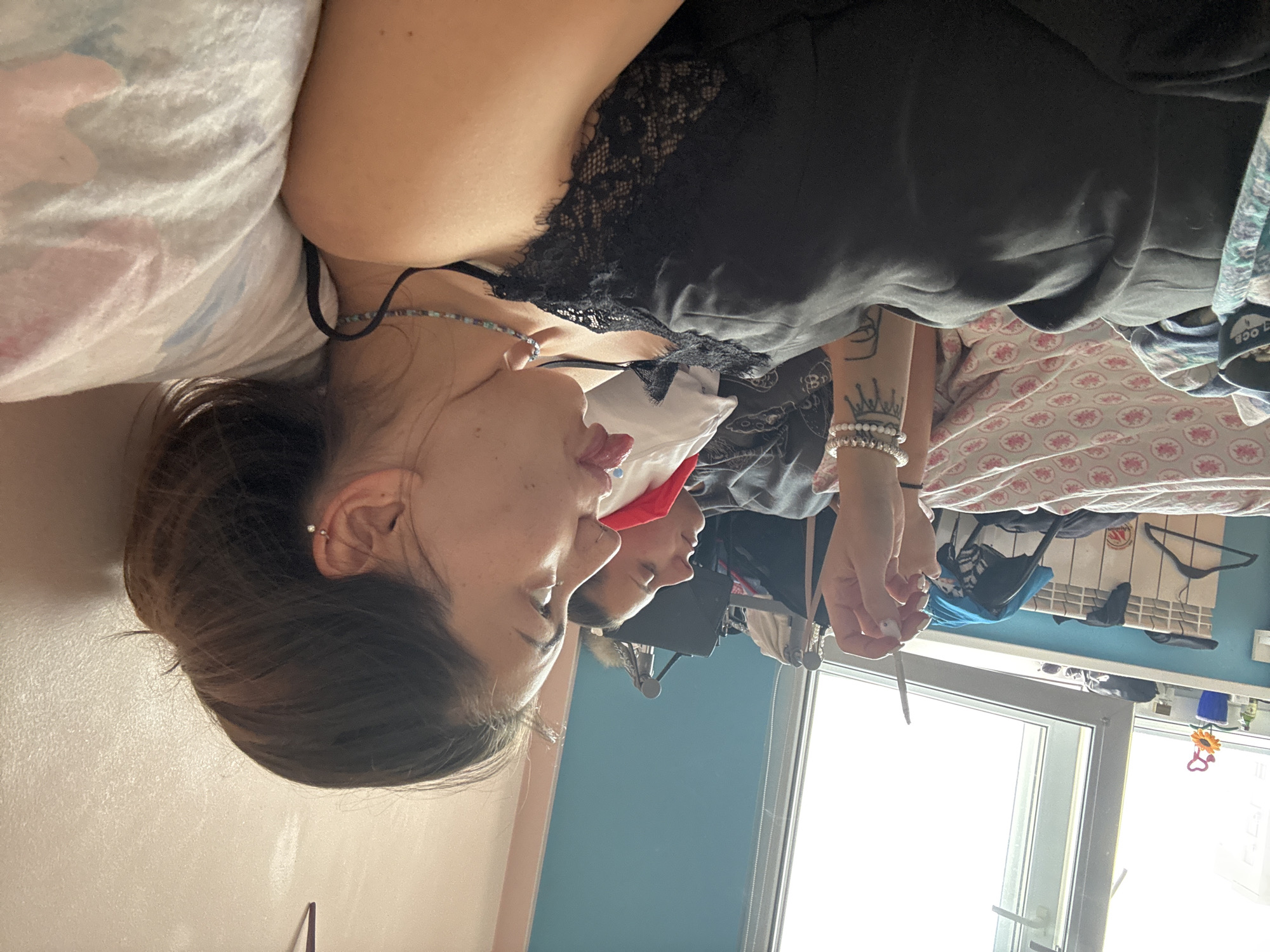 The image depicts two young women in what appears to be a bedroom. The woman in the foreground is lying on her side, facing right, with her tongue sticking out. She is wearing a black lace-trimmed camisole and a beaded necklace. Her hair is dark and tied back. Her right arm is visible, adorned with bracelets and a tattoo. To her left, another woman is lying down, her eyes closed and facing upwards. She is wearing a red top. The room has a large window with natural light streaming in, illuminating a radiator and some clothing items, including a blue garment and hangers, on or near the radiator. A wall with a pale pinkish hue is visible behind the women. The overall impression is that of a casual, indoor setting. There is no specific information in the image to indicate the location is Sofia, Bulgaria.