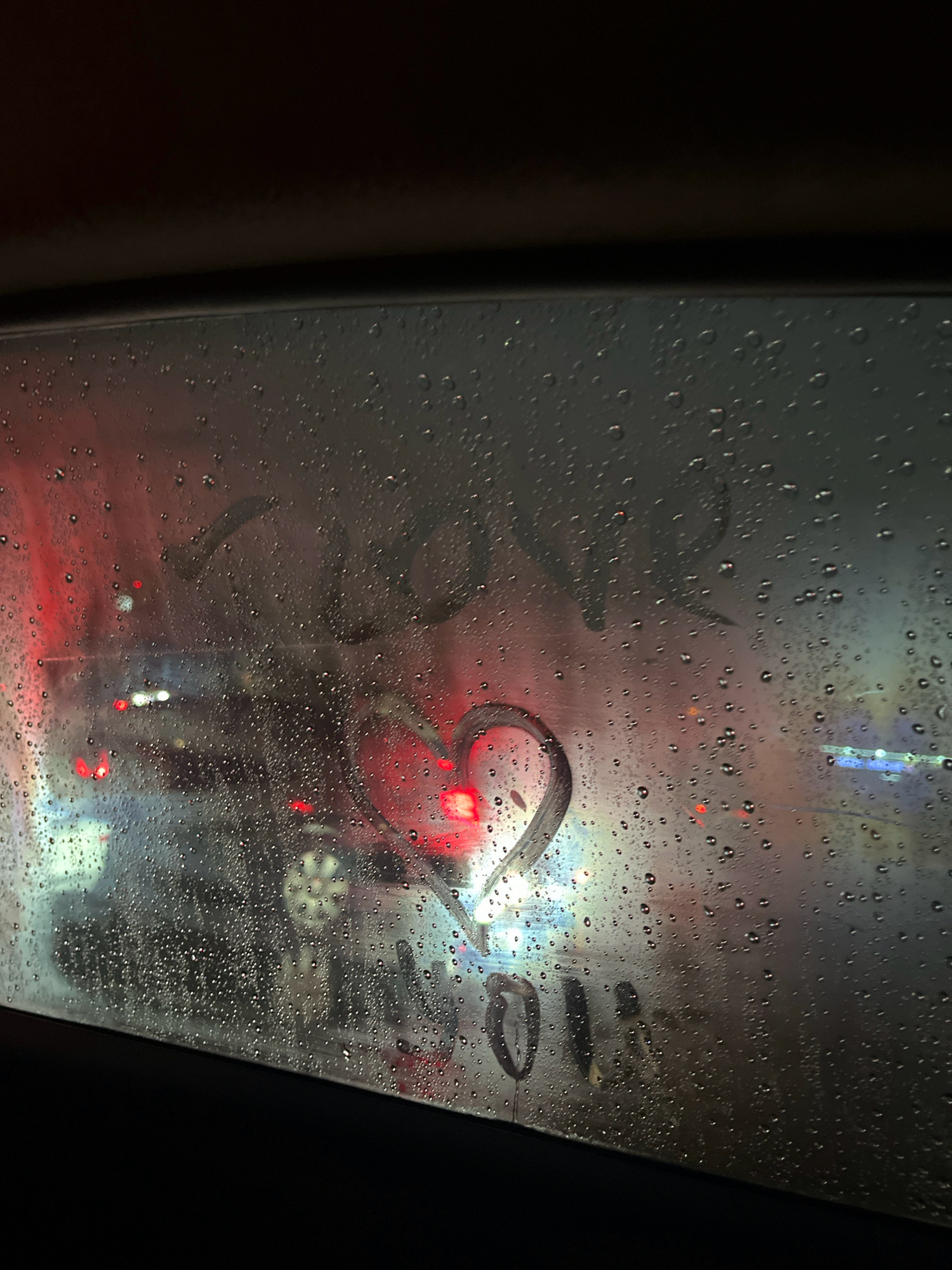 A close-up shot captures a car window fogged with condensation, through which a street scene is visible. Red taillights of traffic ahead are blurred by the water droplets, suggesting a nighttime or rainy setting. A heart shape and some indistinct writing are drawn onto the fogged glass. The environment appears to be an urban area, possibly a traffic jam or a car wash, given the presence of other vehicles and the overall ambiance.