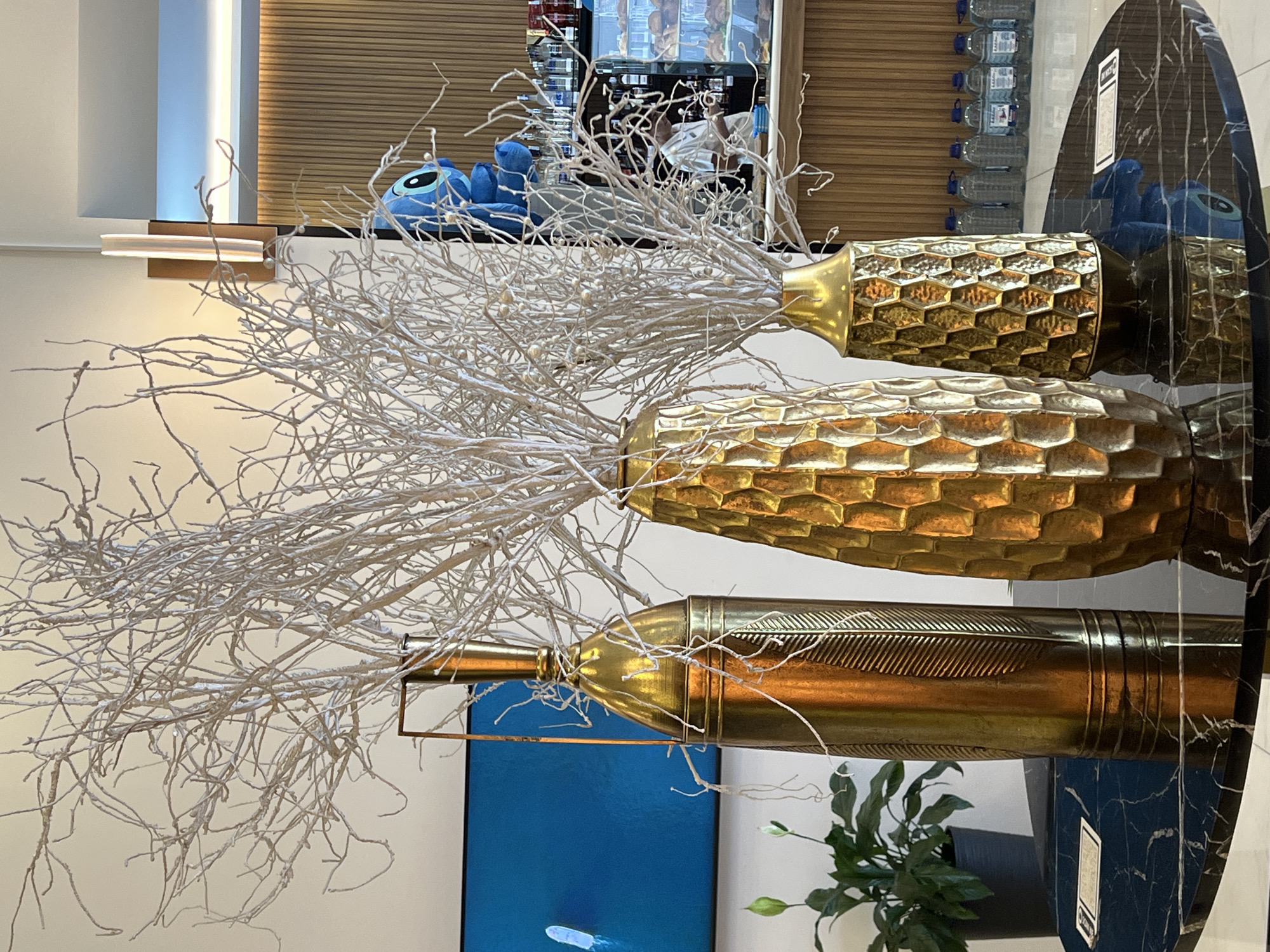 A still life arrangement of three ornate gold vases filled with dried, white, twig-like branches sits on a dark, polished marble-topped table. The vase on the left is tall and cylindrical with a leaf-like etched pattern. The middle vase is wider with a honeycomb-textured surface. The vase on the right is smaller and also features a honeycomb texture. Behind the vases, a blue plush toy, resembling the character Stitch, is partially visible on a shelf. Further in the background, a refrigerated display case contains pastries, and rows of bottled water are arranged on a counter. A person in a light-colored shirt can be seen behind the counter. A flat-screen television displaying a blue image is mounted on the wall to the left. The overall scene appears to be indoors, likely in a cafe or hospitality setting, possibly in Batumi, Georgia, given the context of the request.