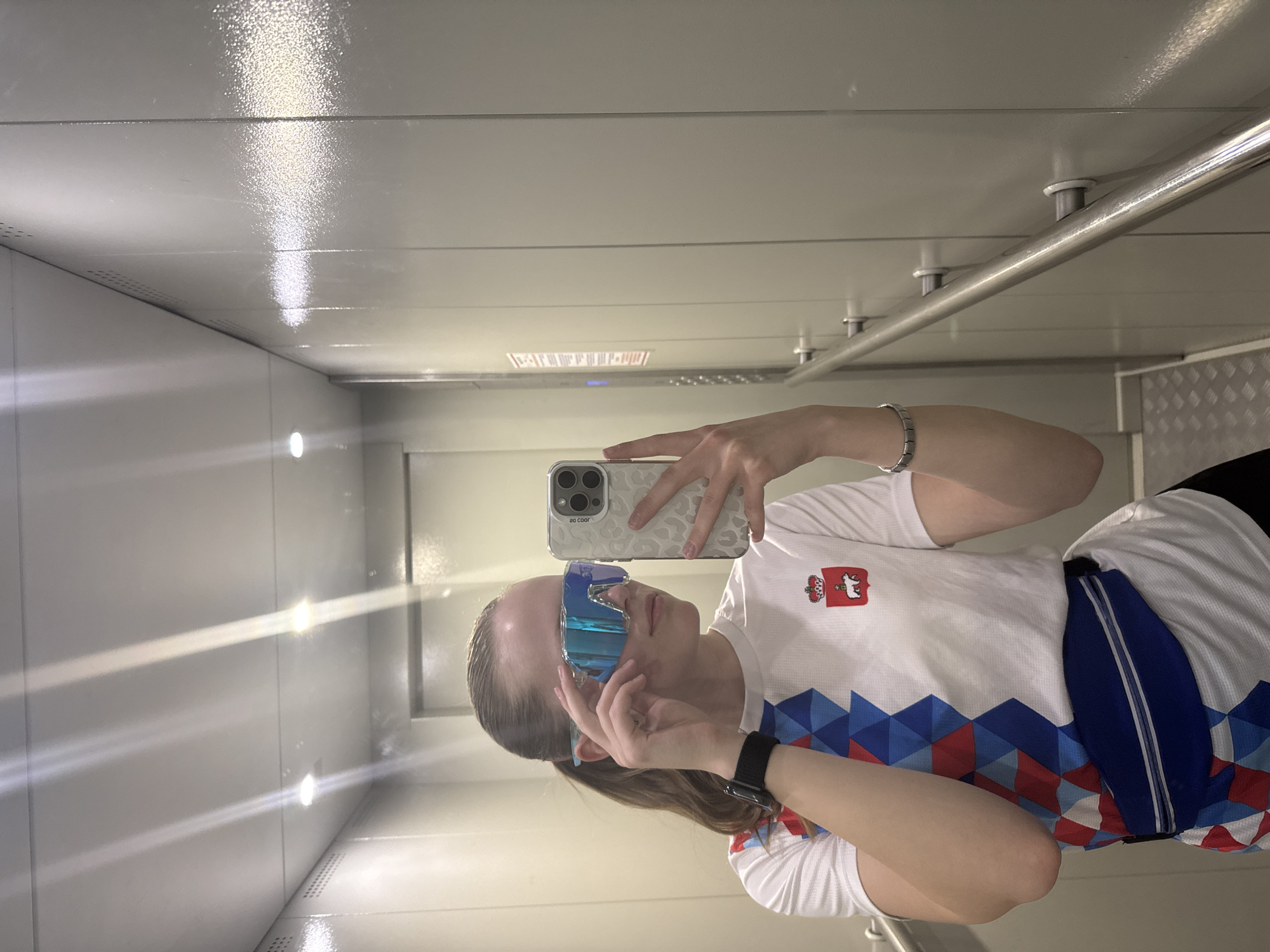 A selfie image shows a fair-skinned young woman inside an elevator in Chaikovsky, Russia. She holds a smartphone in her right hand, angled to capture her reflection in the mirrored elevator wall. She wears oversized, blue-rimmed sunglasses and a white short-sleeved shirt adorned with a red and white crest on the upper left chest and a blue geometric pattern across the lower front. She is also wearing a black smartwatch. The elevator's interior is predominantly light gray, with a shiny, reflective ceiling and a horizontal grab bar along the upper wall. Light fixtures are visible on the side wall.