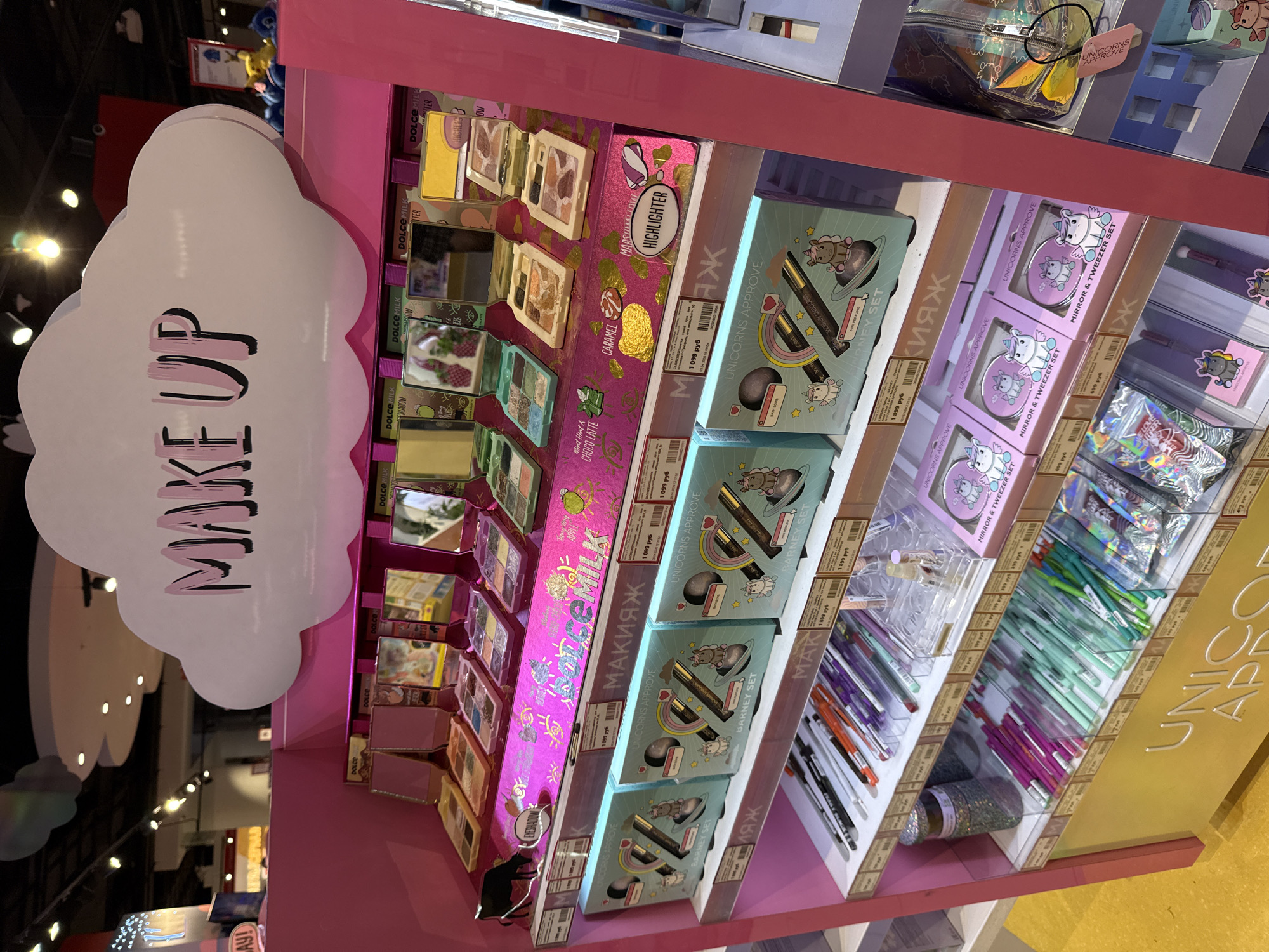 The image displays a brightly lit, multi-tiered retail merchandising unit located within a store in Moscow, Russia. The unit features a pink structural frame, white shelving surfaces, and a yellow base.
At the top, a prominent cloud-shaped sign, rendered in white with pink and black lettering, displays the text "MAKE UP."
The uppermost shelf showcases multiple open compact cases from the "DOLCE MILK" brand, each containing eyeshadow palettes or highlighters. These compacts feature integrated mirrors. Visible product identifiers include "EYESHADOW," "HIGHLIGHER," "CHOCLATE," "CARAMEL," and "MARSHMALLOW." A small black bovine figurine is positioned on the left side of this shelf. White price labels, displaying values such as "1099 руб," are affixed beneath these products.
The second shelf from the top presents three identical light blue gift sets labeled "UNICORNS APPROVE" and "BARNEY SET." Each box depicts a bath bomb, a pen or cosmetic liner, and another small item, accompanied by cartoon unicorn illustrations. Price labels of "1899 руб" are visible for these sets. The Russian word "МАКИЯЖ" (Makeup) is written in white on the pink vertical panel behind this shelf.
The third shelf contains multiple rows of pens or similar writing instruments, organized by color, including black, grey, orange, purple, green, and pink variants.
The bottom shelf displays additional "UNICORNS APPROVE" merchandise, specifically several pink square boxes marked "MIRROR & TWEEZER SET," each featuring a unicorn graphic. A cylindrical container filled with glitter is also present on this shelf. The yellow base of the display unit shows partial white lettering that reads "UNICO APP," likely an abbreviation for "UNICORNS APPROVE."
The background indicates a larger retail environment, with blurred views of other product displays and overhead lighting fixtures. All displayed prices are denoted in Russian Rubles (руб). No individuals are visible in the scene.
