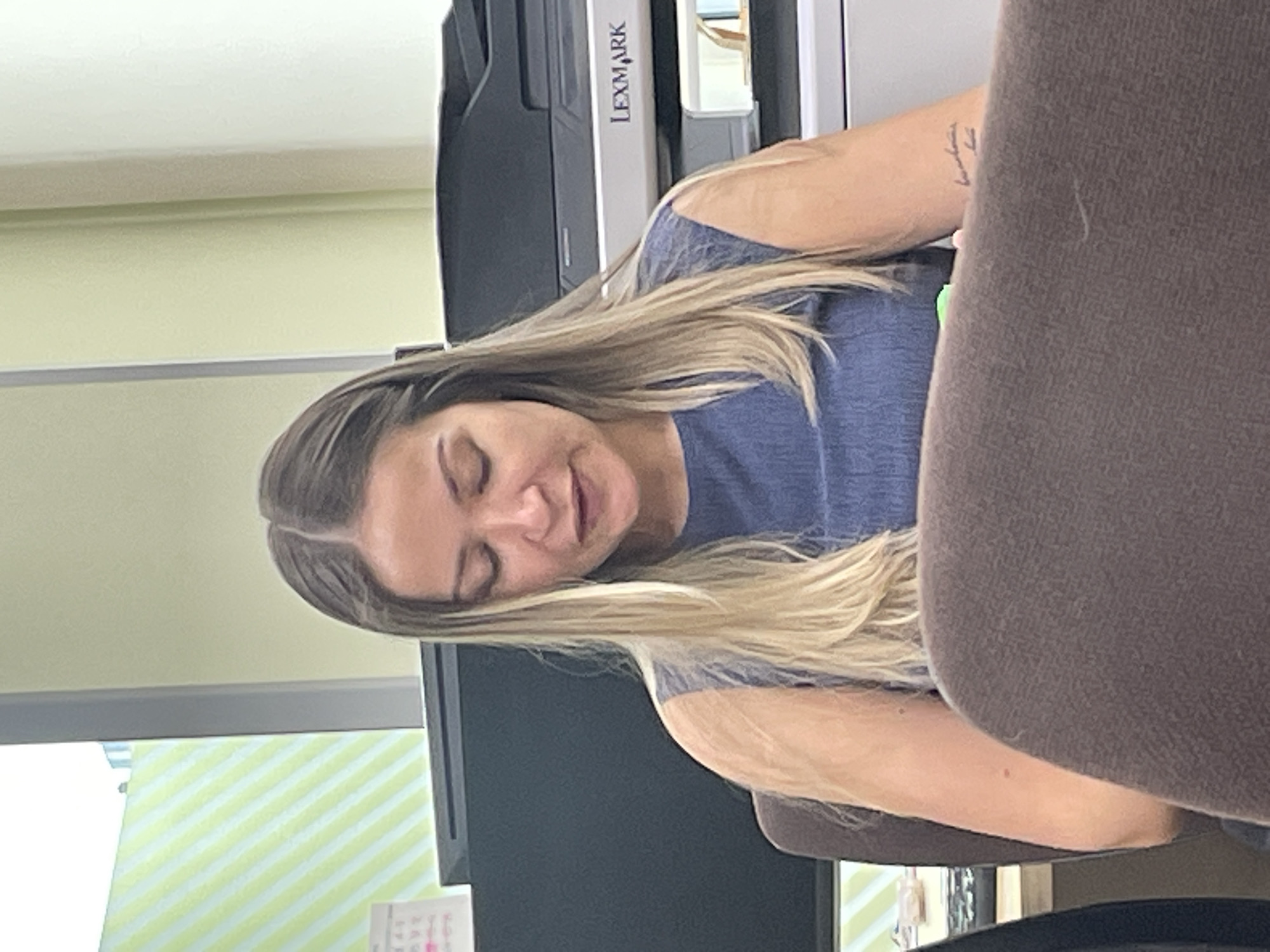 This image, set in Krasnoyarsk, Russia, captures a candid indoor scene of a woman. She has long, straight hair with a light brown base transitioning to blonde ends, and is wearing a dark blue short-sleeved top. Her head is tilted, and her eyes are closed or partially closed, with her mouth slightly open, suggesting a moment of rest, reflection, or perhaps tiredness.
The setting appears to be an office or workspace. In the background, a large, dark-colored Lexmark multifunction printer or copier is prominently visible, bearing the "LEXMARK" brand name. To the left of the woman, a portion of a computer monitor or screen is visible, with a small desk item (possibly a calendar) underneath it. The walls are painted a light green or pale yellow hue. On the far left, there is a source of light, possibly a window, showing light green and white diagonal stripes, which could be from blinds or a pattern on an object.
A dark brown chair, likely where the woman is seated, partially frames the right side of the image. A tattoo with unidentifiable script is visible on her right forearm. The lighting indicates an indoor daytime setting. The woman's relaxed posture and expression convey a sense of quiet or introspection.