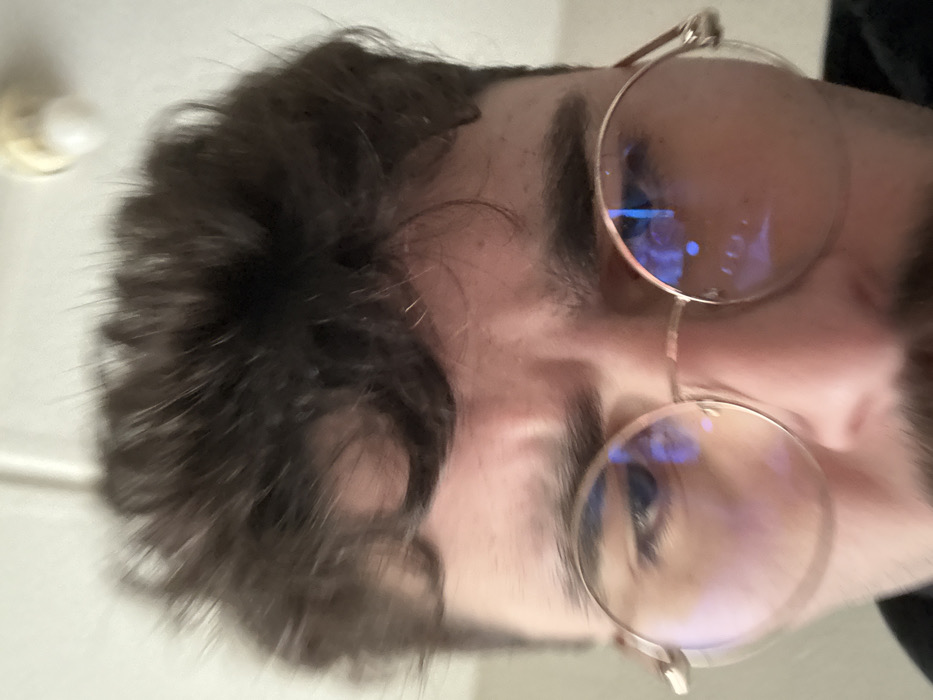 An extreme close-up, low-angle shot features the upper face of a male individual, likely a young adult. The subject has dark, curly hair falling over his forehead, thick dark eyebrows, and visible horizontal creases on his brow, suggesting a furrowed expression. He wears round, gold-framed glasses; the lenses exhibit distinct blue light reflections, possibly from a digital screen or artificial light source. His eyes appear dark behind the lenses, and the bridge of his nose is visible along with a dark mustache. The background is uniformly light-colored and out of focus, revealing a portion of a ceiling with a blurry, cream-colored fixture, possibly a light bulb socket, in the top right. The environment is an indoor setting, but the specific location within the city of Rennes, France, cannot be determined from the visual information provided. No direct interactions are depicted.