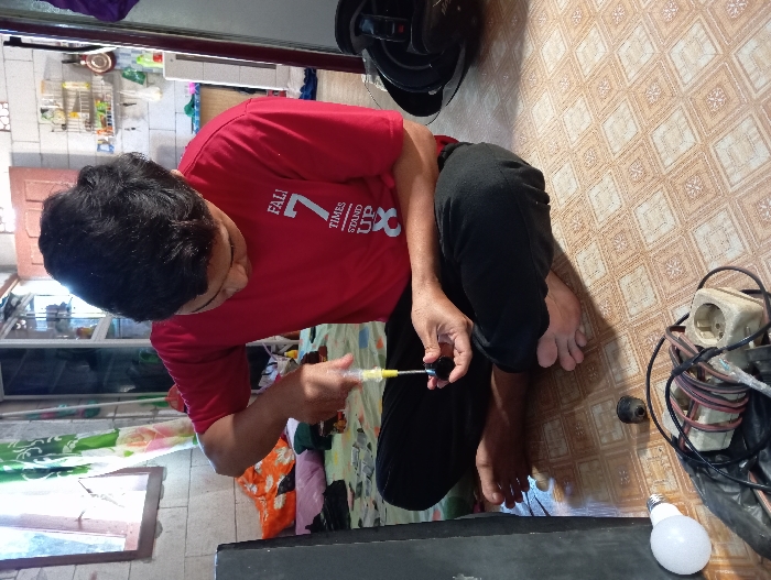 A male individual, dressed in a red short-sleeved t-shirt with white text and black long pants, is seated cross-legged and barefoot on a light brown, geometrically patterned tiled floor. He is actively engaged in repairing a small, black electrical component, holding it in his left hand while using a yellow-handled screwdriver in his right hand to manipulate its internal mechanism. His attention is focused downward on the task. To his left on the floor are a white light bulb, a tangled bundle of electrical wires, a beige multi-socket power strip, and a black plastic bag containing additional items. A large, dark rectangular object is partially visible at the extreme left. The background features a window with wooden framing and green and white patterned curtains on the left, an unmade bed or pile of textiles behind the individual, and a rear wall with white rectangular tiles and a shelving unit displaying various bottles and containers. A dark object, possibly a helmet, rests on the floor to the individual's right. The scene is illuminated by natural light entering from the left. This event occurs in Baron, Indonesia.