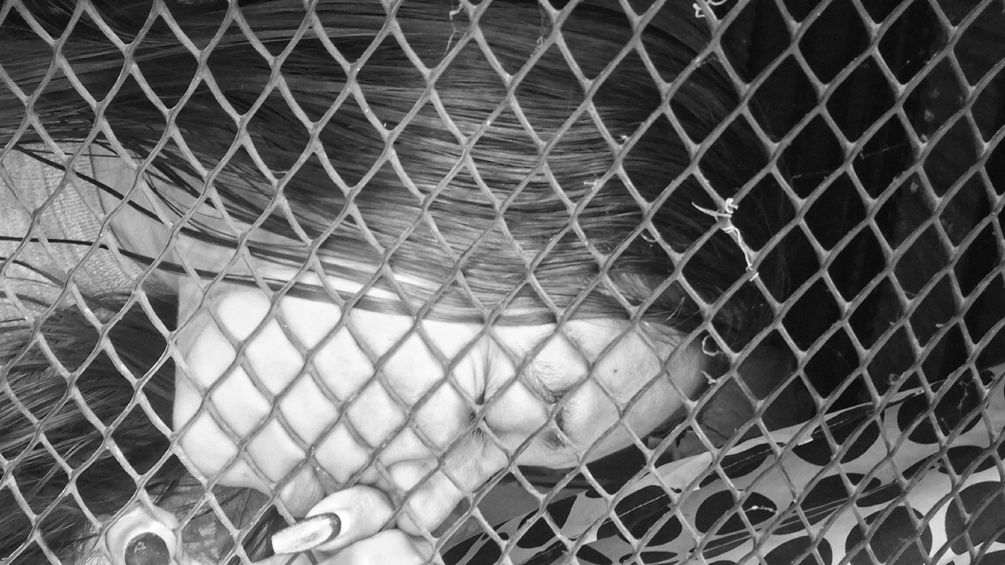 This is a black and white, close-up, high-angle shot of a person, viewed through a diamond-patterned fence. The subject's face is largely obscured by their hair, but their cheek and eye are visible, closed. The person appears to be lying down, with their head resting on a patterned surface, possibly fabric or a blanket, which shows a repeating circular design. Their hair is dark and extends across the frame. One hand is visible in the lower left corner, with painted fingernails. The lighting suggests it is daytime, but there are no clear indications of weather or specific location cues beyond the fence. The overall mood is somber and possibly indicative of distress or weariness. No text is visible in the image.