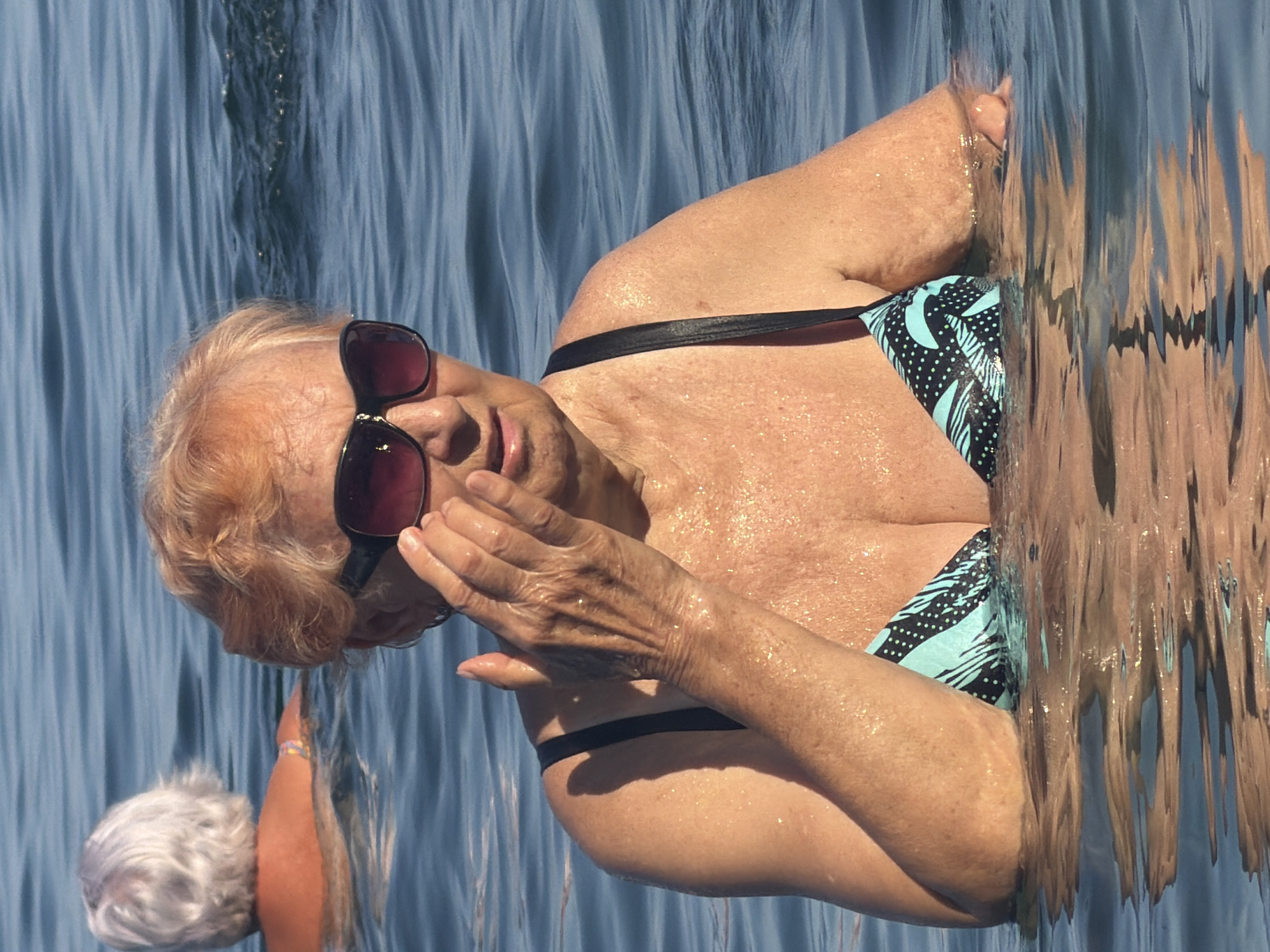 An elderly woman with blonde hair and sunglasses is swimming in the sea near Partenit, Ukraine. She is wearing a black and blue patterned bikini top. Her skin is wet and glistening in the sun. Another elderly person with white hair is partially visible in the background, also in the water. The water is blue and calm with gentle ripples.